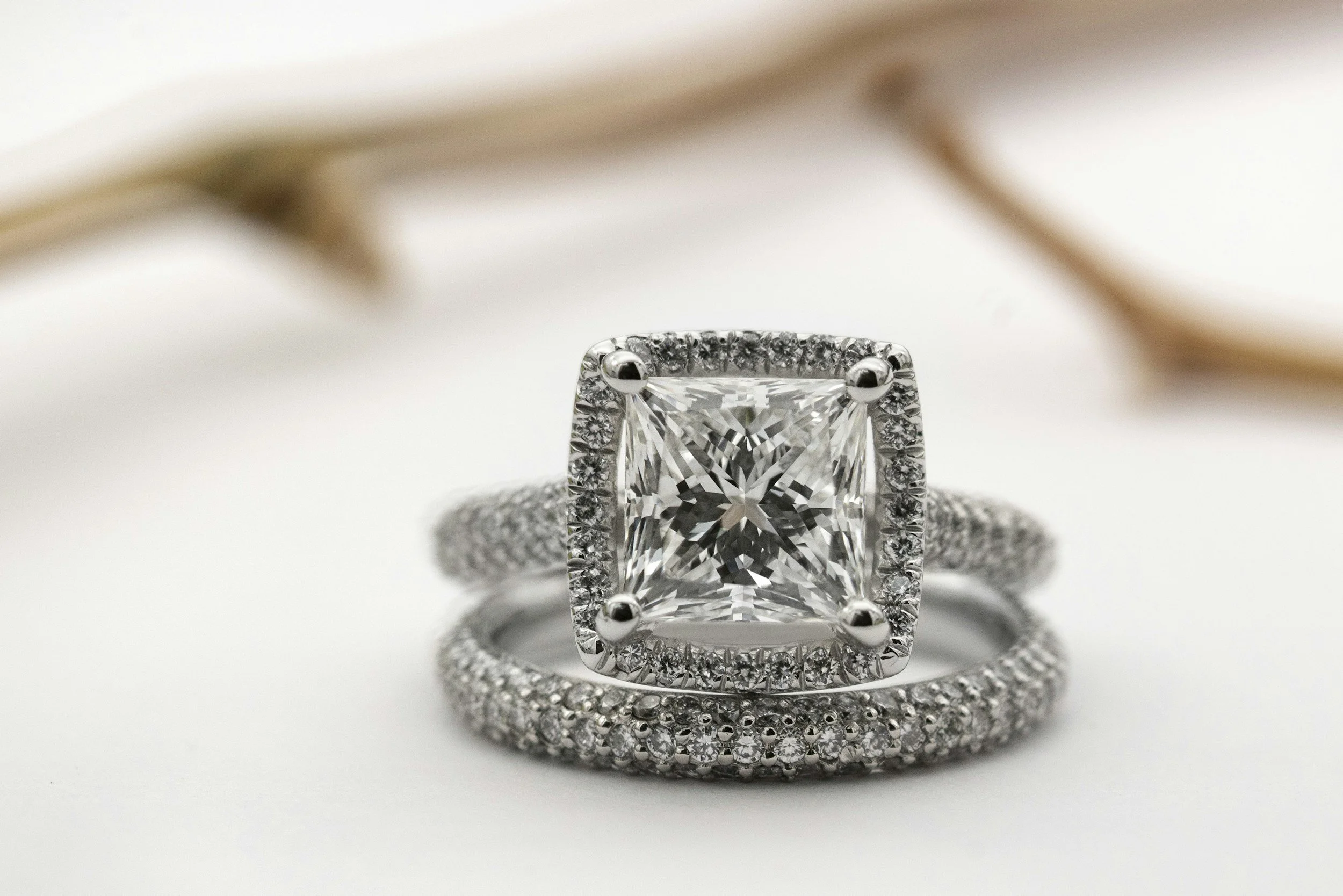 Close-up of an elegant engagement ring with a large square-cut diamond surrounded by smaller diamonds, set on a silver band with additional diamonds, against a soft white background with blurred elements in the background.