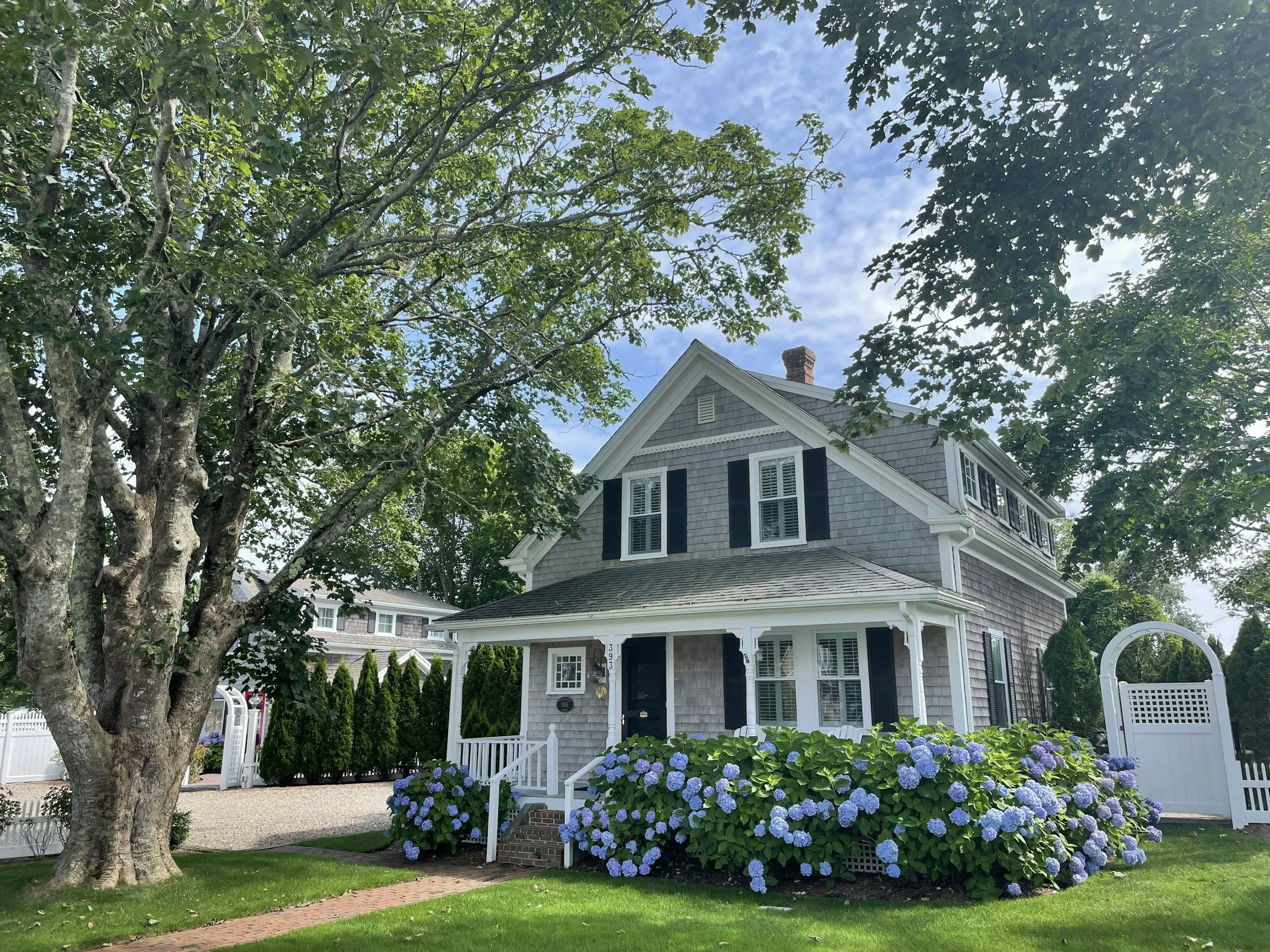 A two-story house with gray shingles, white trim, and black shutters, surrounded by lush green trees and blooming hydrangeas, with a white picket fence and gate.