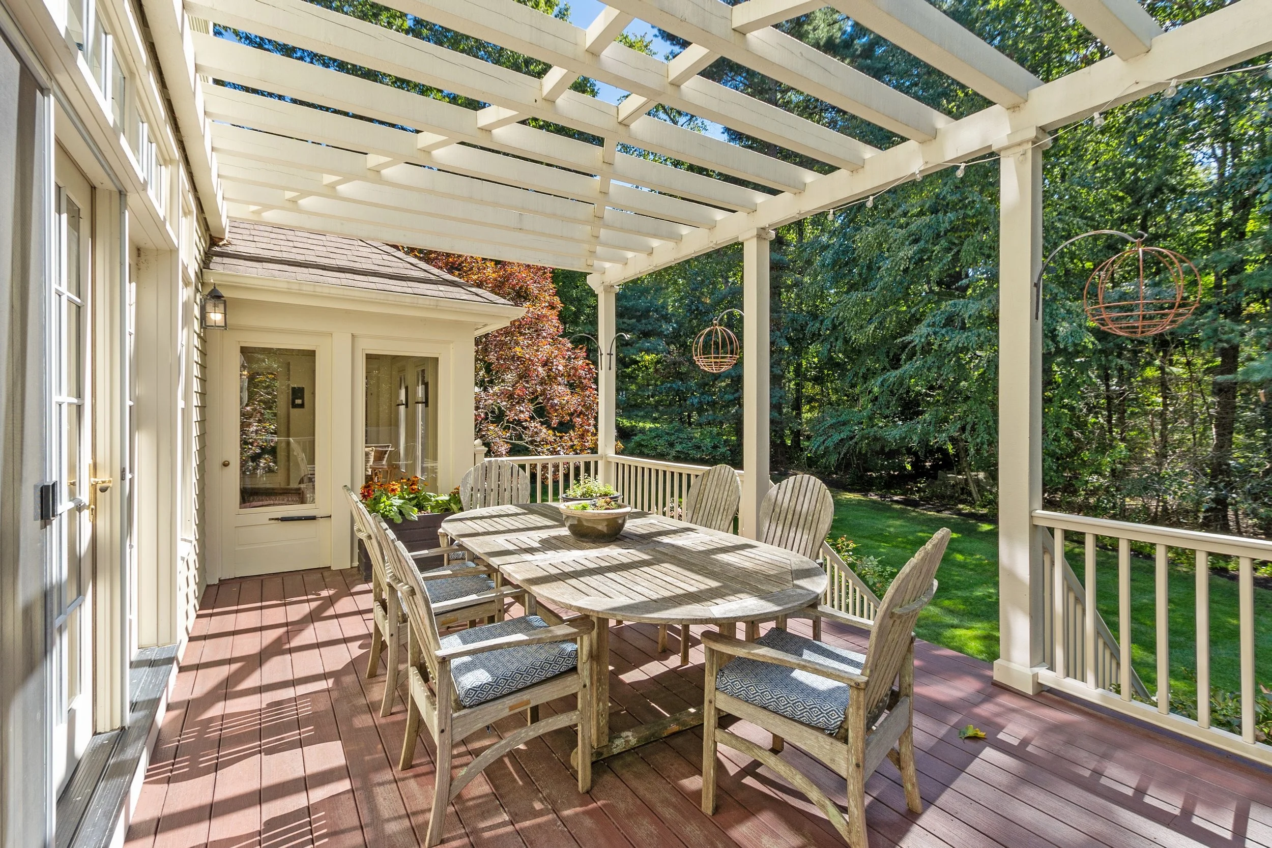 Outdoor patio with wooden dining table, chairs, and hanging spherical lanterns surrounded by lush trees and greenery