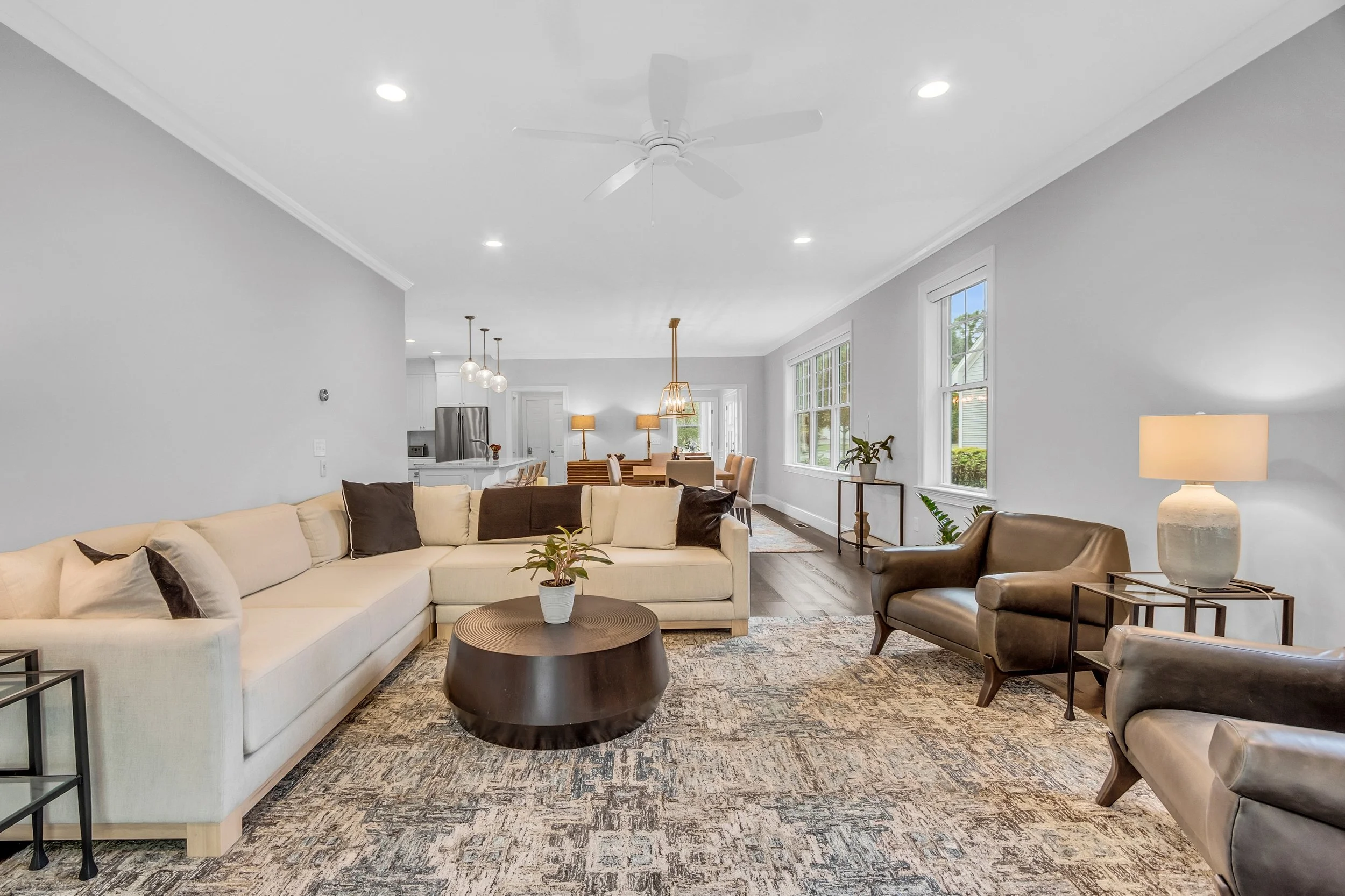 Living room with white sofa, brown leather armchairs, wooden coffee table with plant, patterned rug, white walls, large windows, and modern decor. Kitchen and dining area visible in background.
