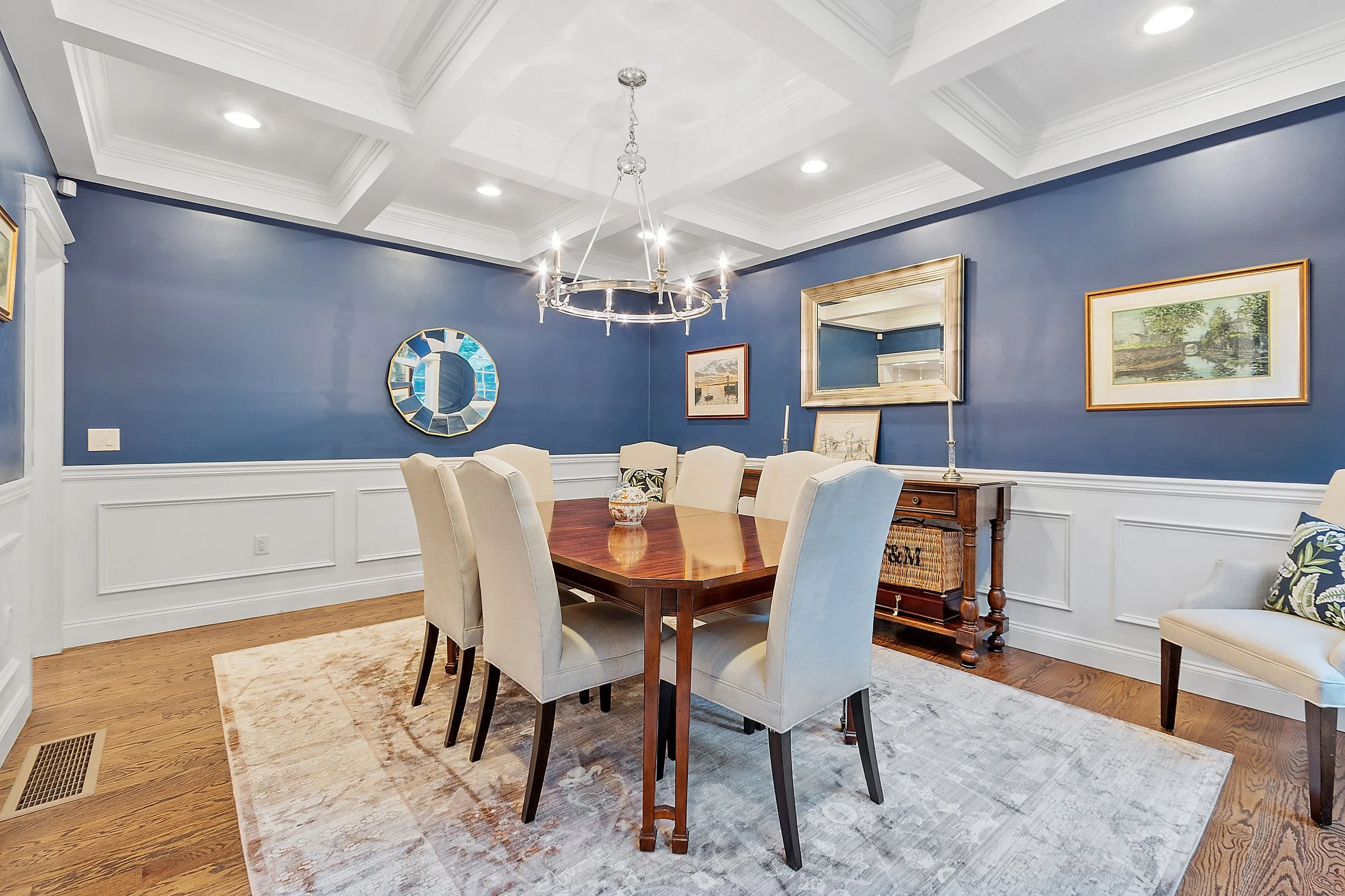 Dining room with blue walls, white wainscoting, a wooden table with beige chairs, a chandelier, framed artwork, a mirror, and a side table.