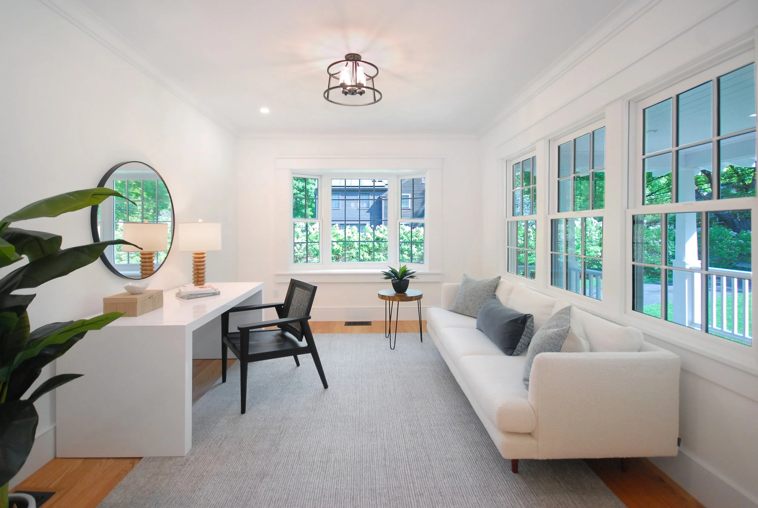 Bright living room with white walls, large windows with greenery outside, a white sofa with gray and black pillows, a small side table with a plant, a black desk chair, a white desk with lamps, a round mirror, a ceiling light fixture, and a light-colored rug on hardwood floors.