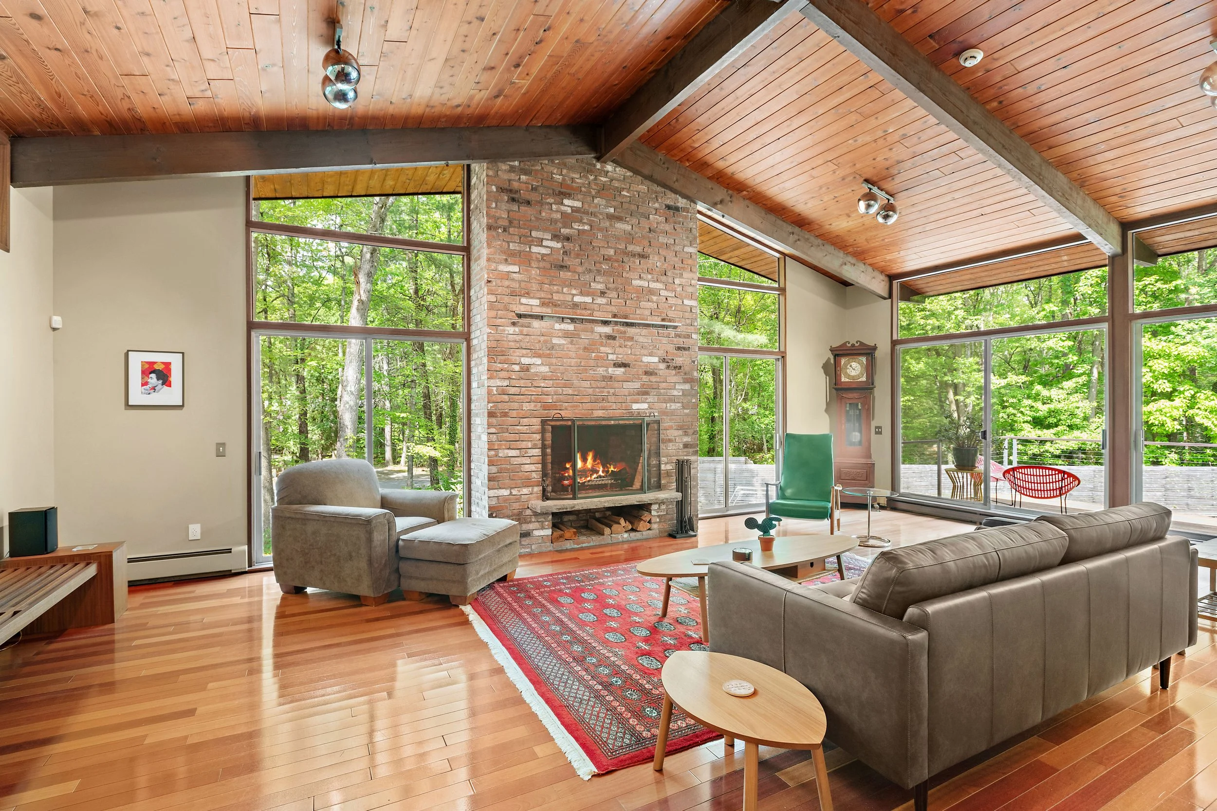 Living room with large windows overlooking a wooded area, a brick fireplace with a fire burning, gray sofa, armchair, wooden coffee table, vintage clock, colorful rug, and wooden ceiling beams.