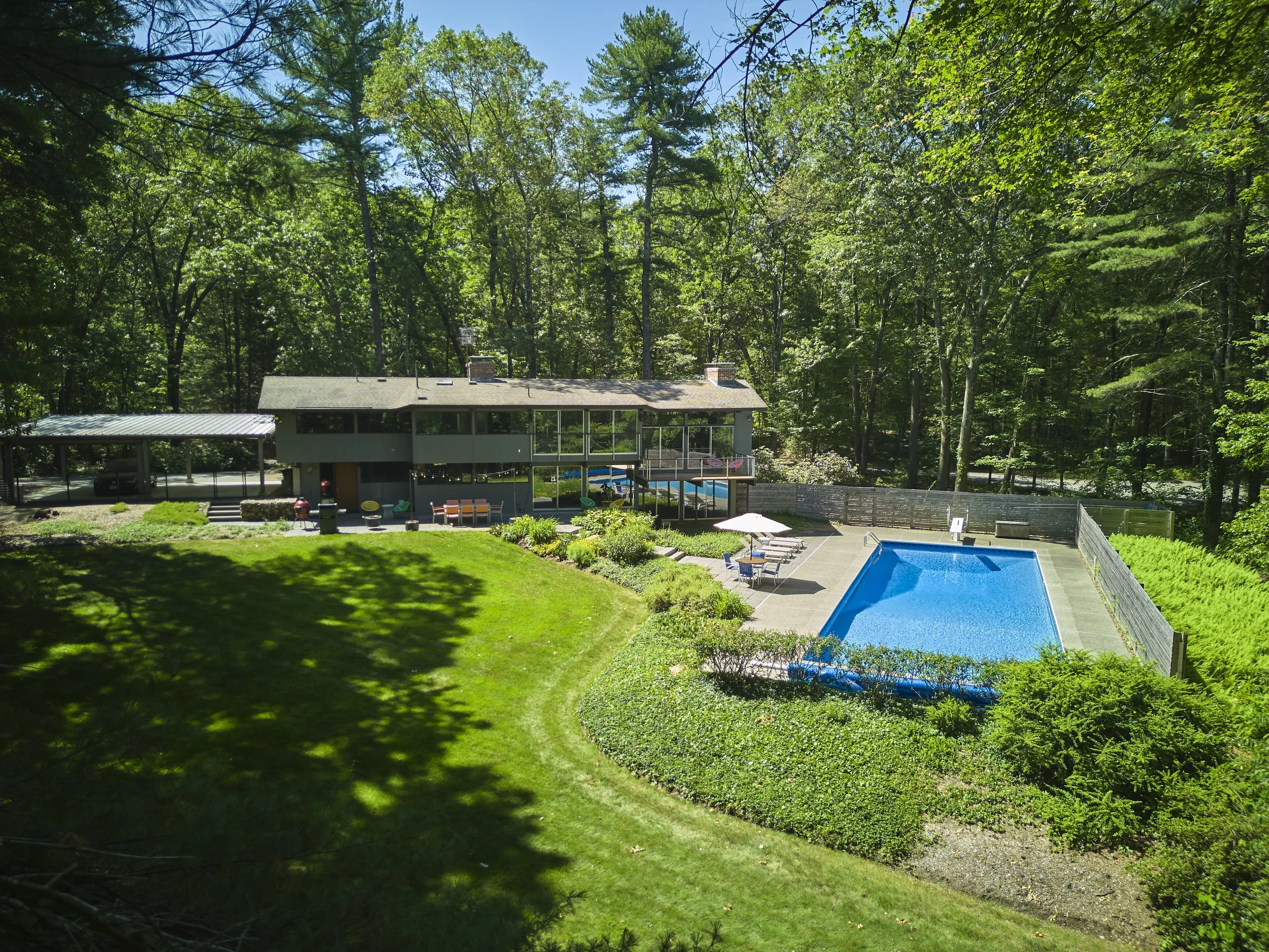 View of a backyard with a swimming pool, patio furniture, a house, and surrounded by trees.