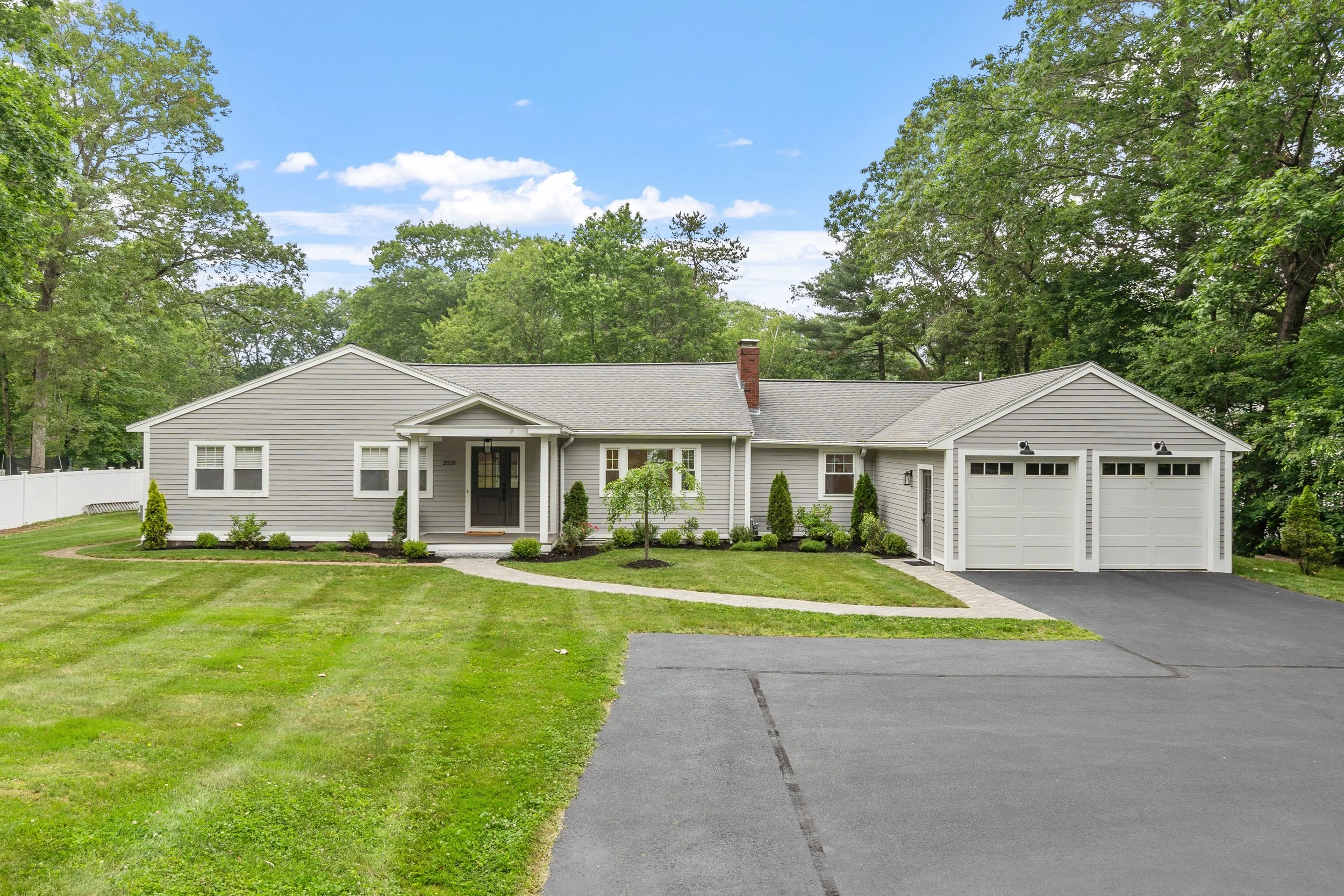 Front view of a single-story house with gray siding, two-car garage, front lawn with small tree and bushes, concrete driveway, surrounded by green trees, blue sky with white clouds