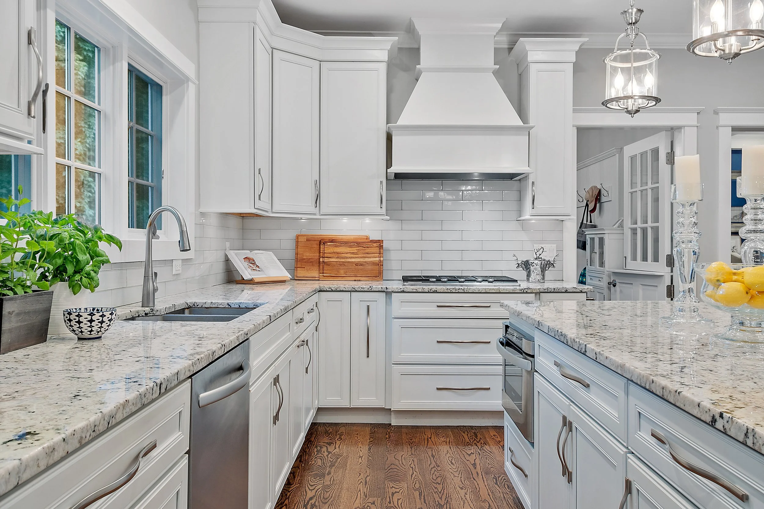 A modern kitchen with white cabinets, granite countertops, a stainless steel dishwasher, and a stove with a range hood. There are windows above the sink, potted plants, and decorative candles on the island.
