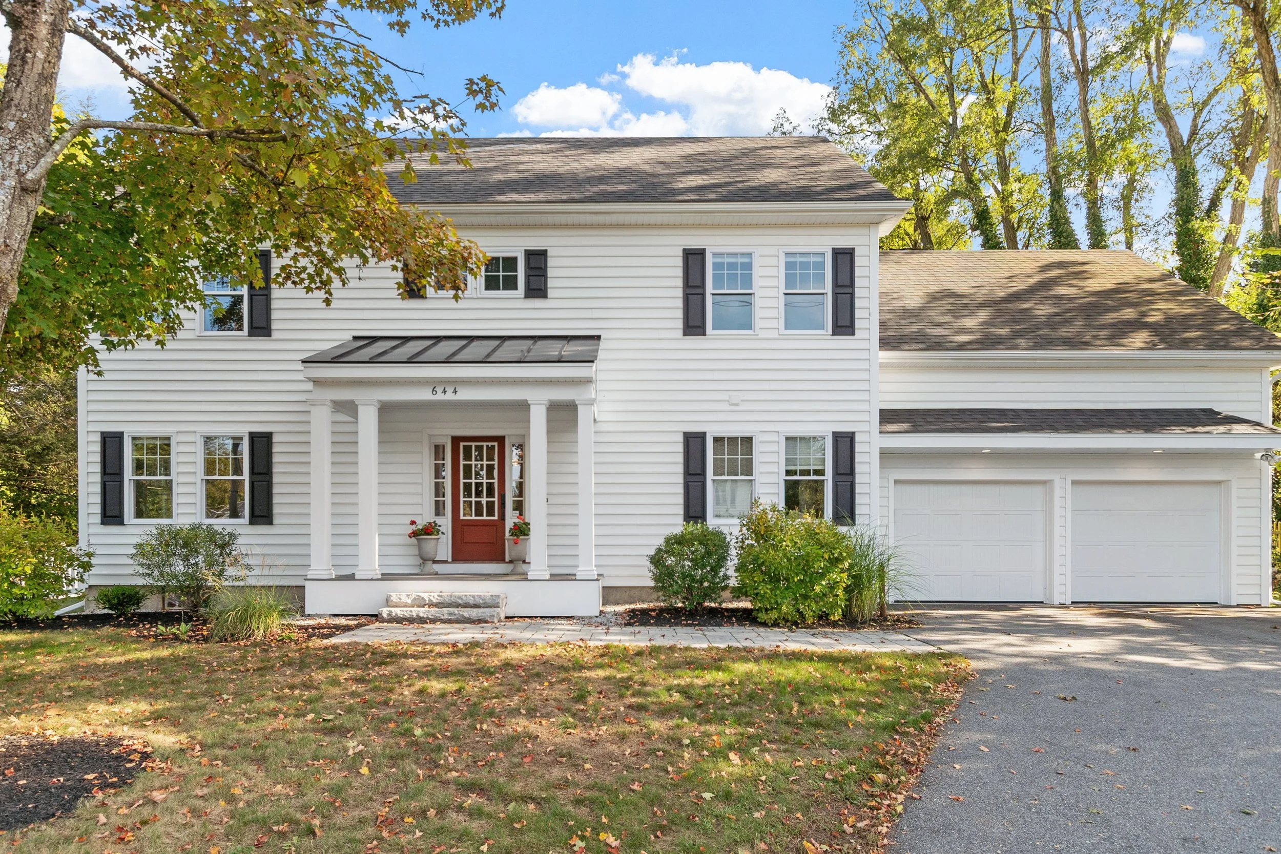 A two-story white house with black shutters, a red front door, and a double garage. The house is surrounded by trees with green leaves and a lawn with patches of fallen leaves.