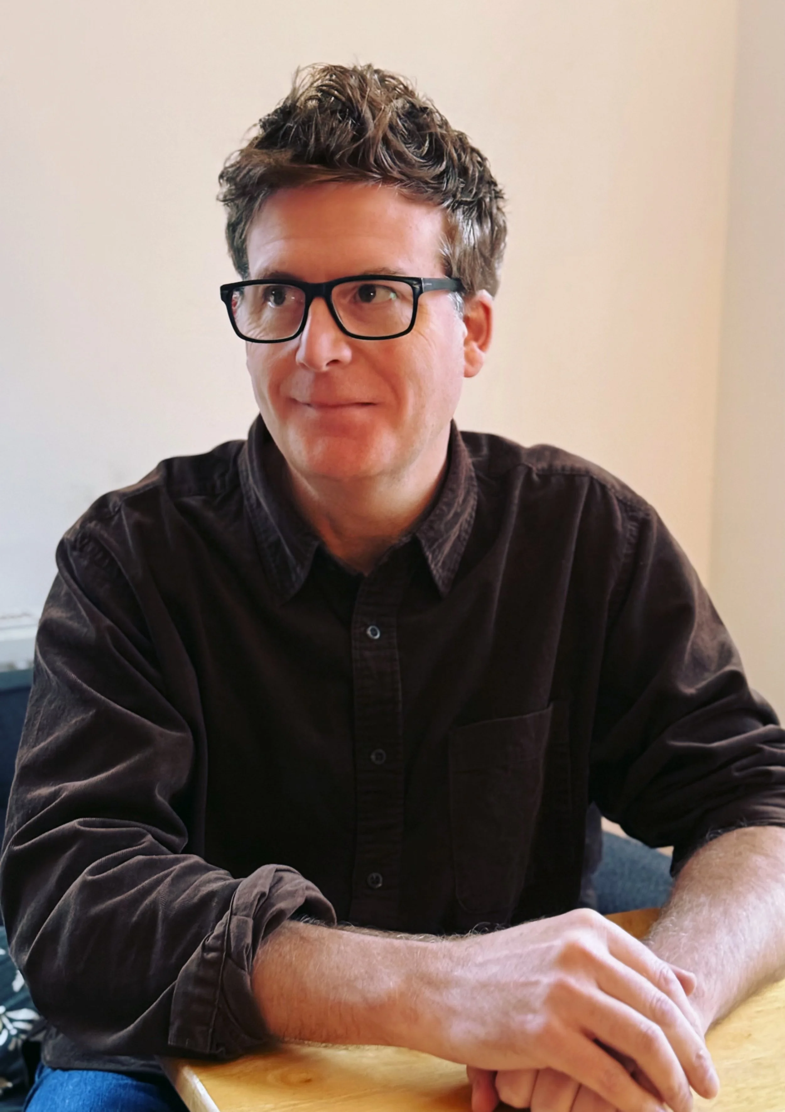 A therapist with brown, wavy hair, black glasses, wearing a dark button-up shirt, sitting at a table.
