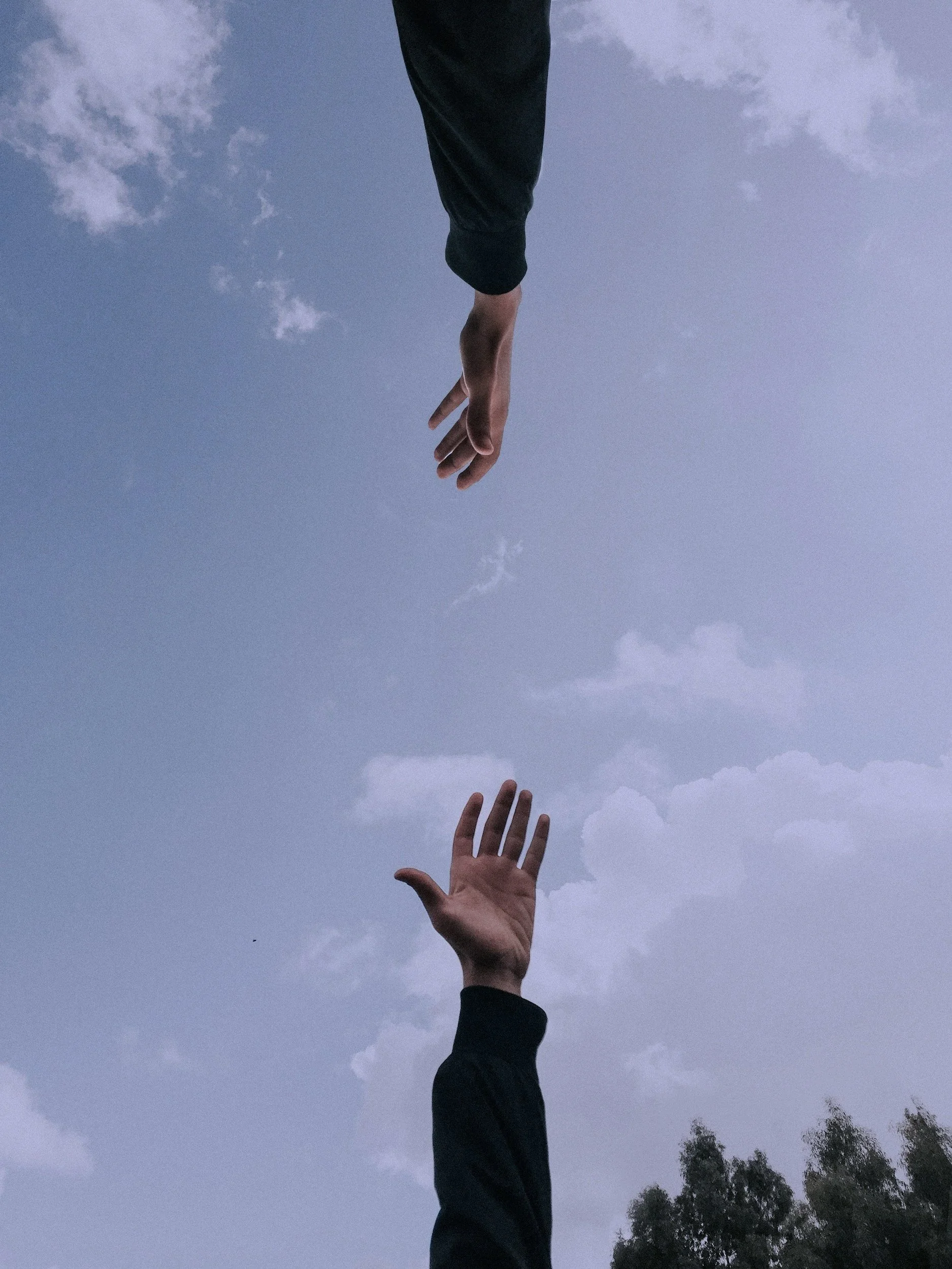 A surreal image showing two hands reaching toward each other against a cloudy sky, one hand from above and the other from below, creating an illusion of connection or attempt to touch.