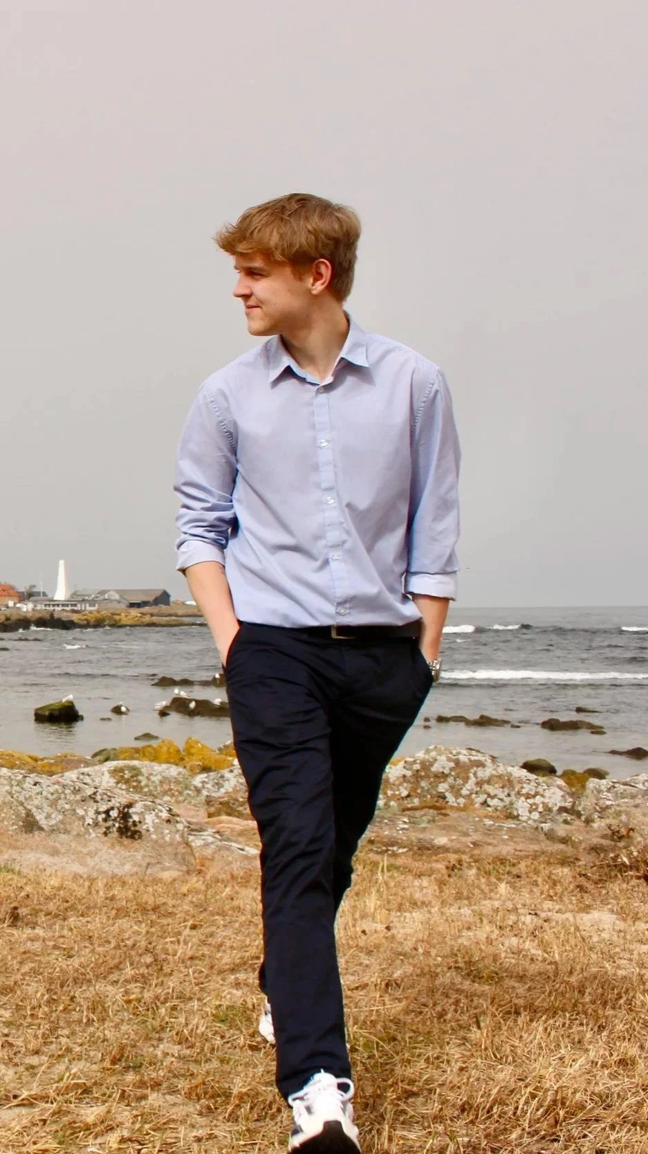 Ung mand går langs en strand med sten og hav i baggrunden, iført let tøj og har hænder i lommer.