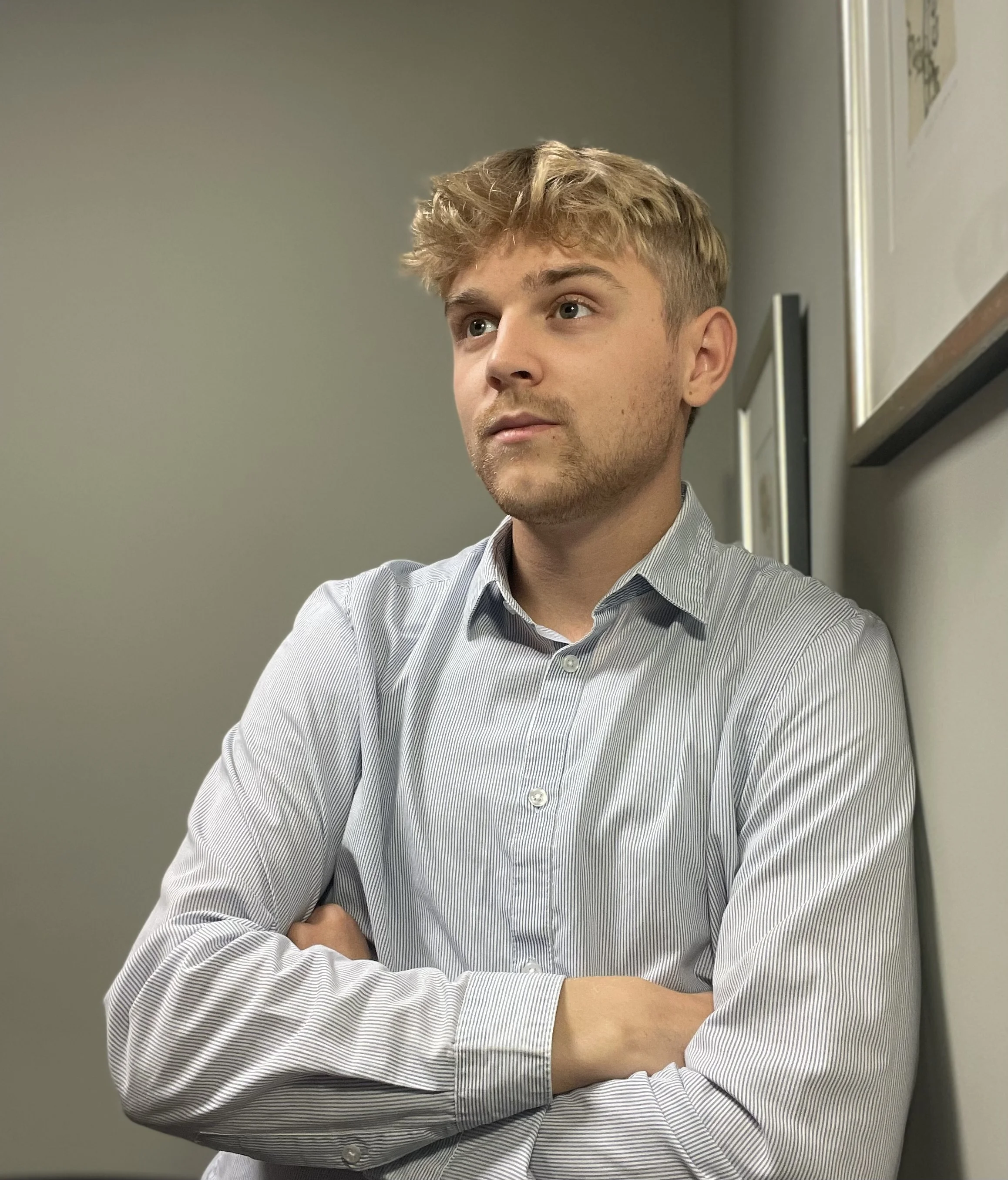 Ung mand med blond, let krøllet hår og skal af, iført stribet skjorte, står med armene korsede ved en væg i et kontor. 