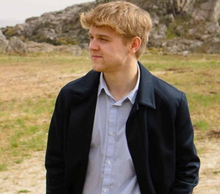 En ung mand med blondt hår iført en hvid skjorte og sort jakke står udendørs foran en klippeformation.