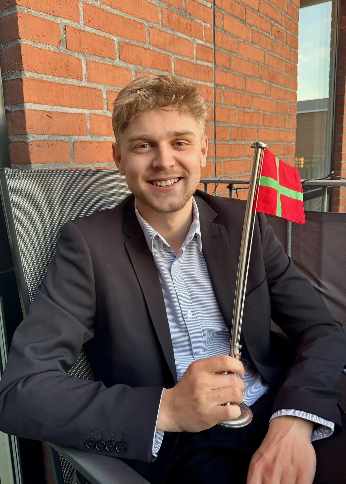 En glad ung mand iført mørk habit sidder i en stol på en balkon med et bornholmsk flag, foran en murstensvæg.