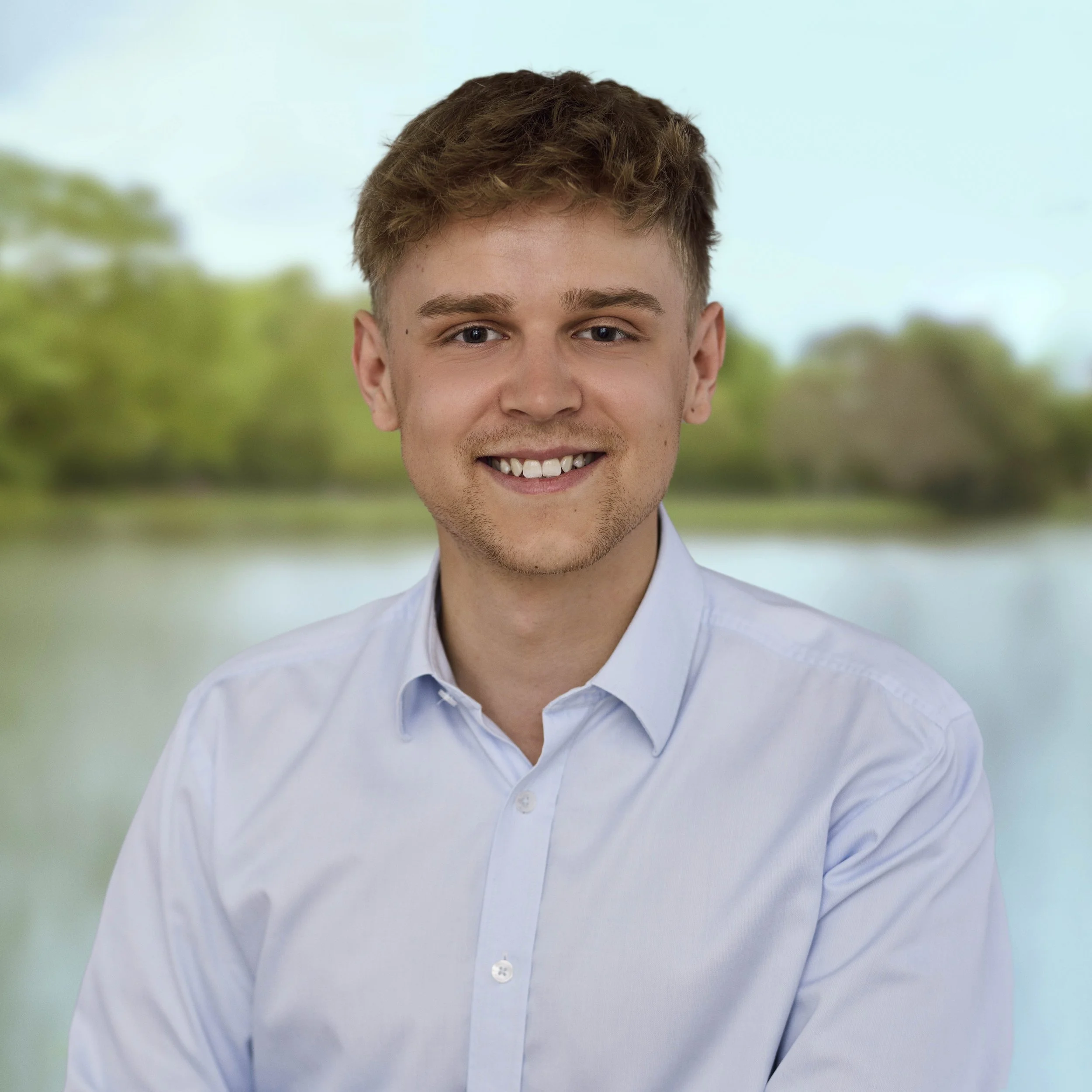 En ung mand med kort lysebrunt hår, iført en hvid skjorte, smiler foran en naturlig baggrund med vand og grønne træer.