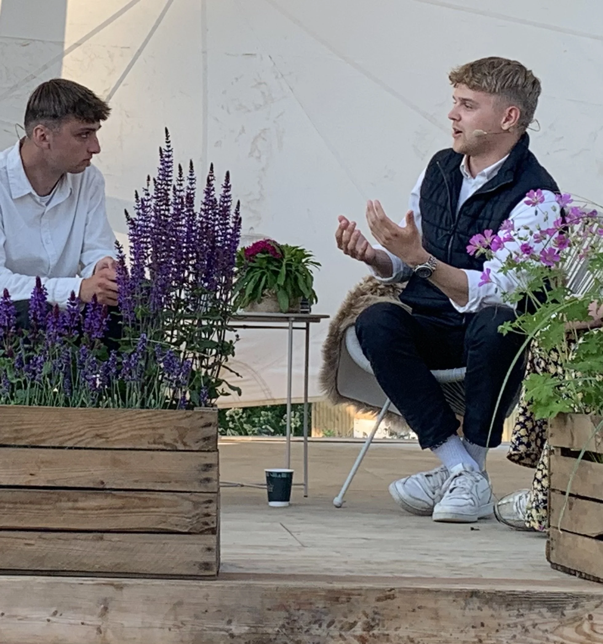 To mænd sidder og taler i en udendørs setting med blomster og potter omkring dem.