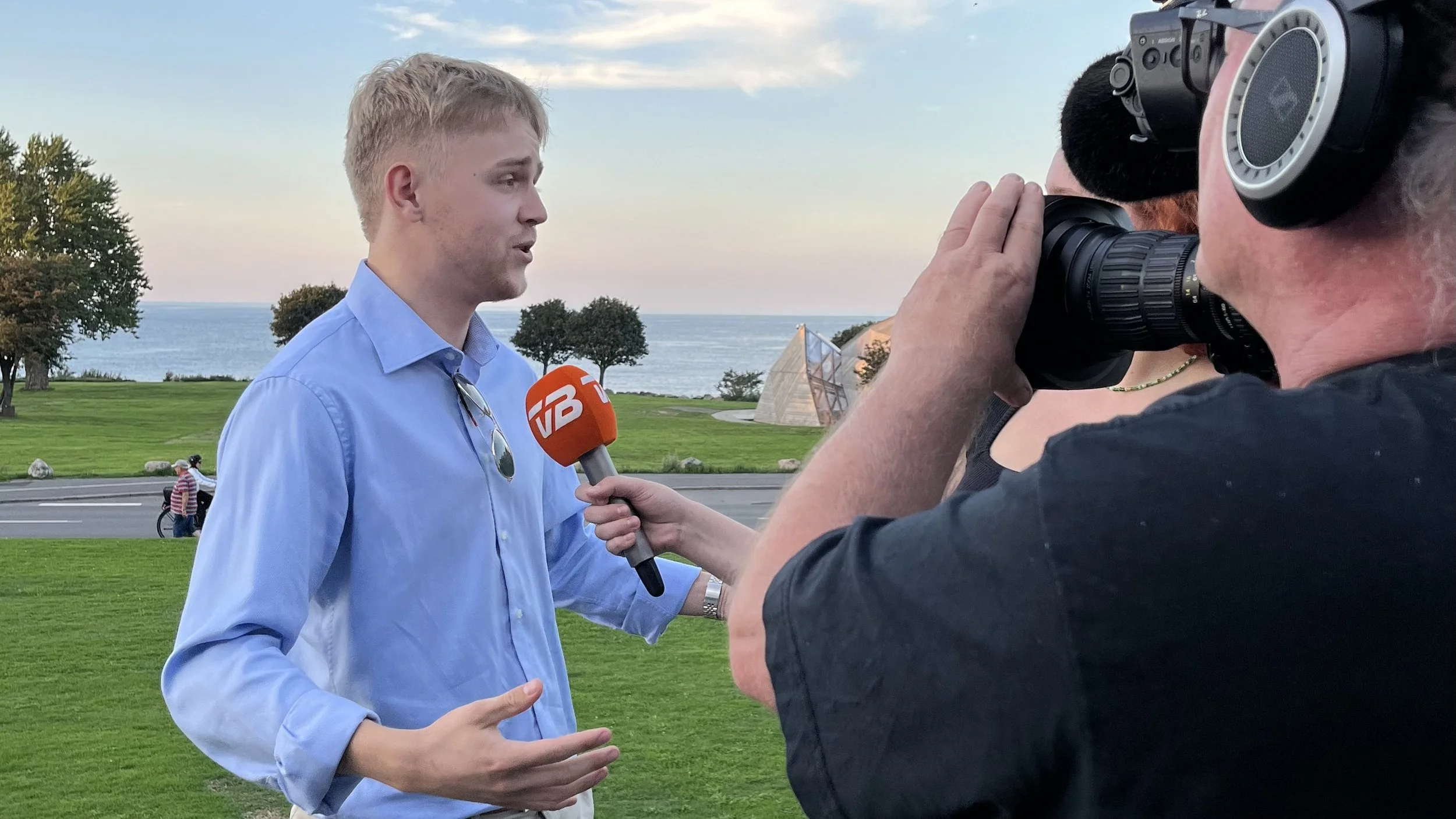 En mand, der bliver interviewet udendørs med et kamera og mikrofon, foran en grøn park med træer, en vej og vand i baggrunden.