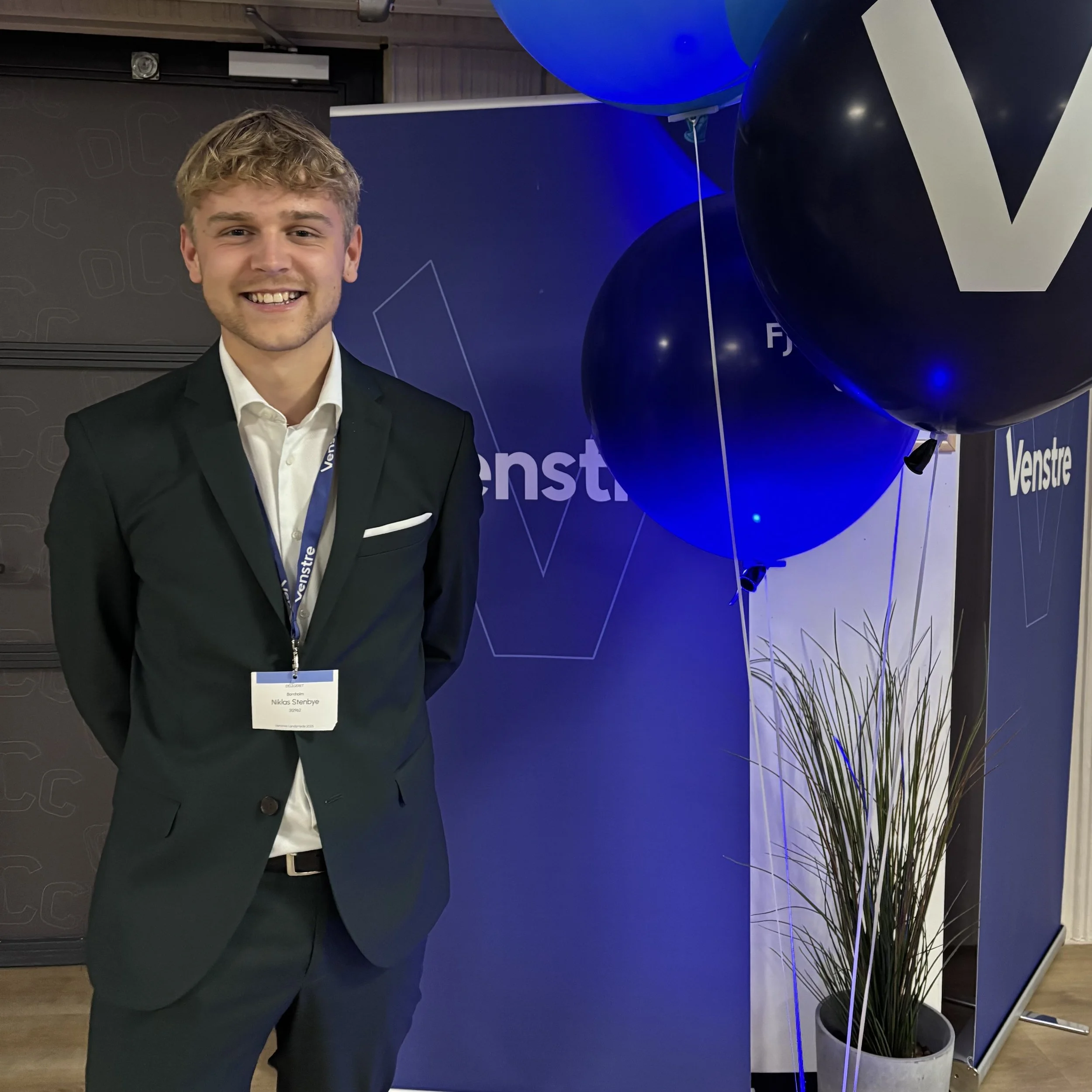 En ung mand i jakkesæt står foran en blå baggrund med teksten "Venstre" og nogle balloner, mens han smiler.