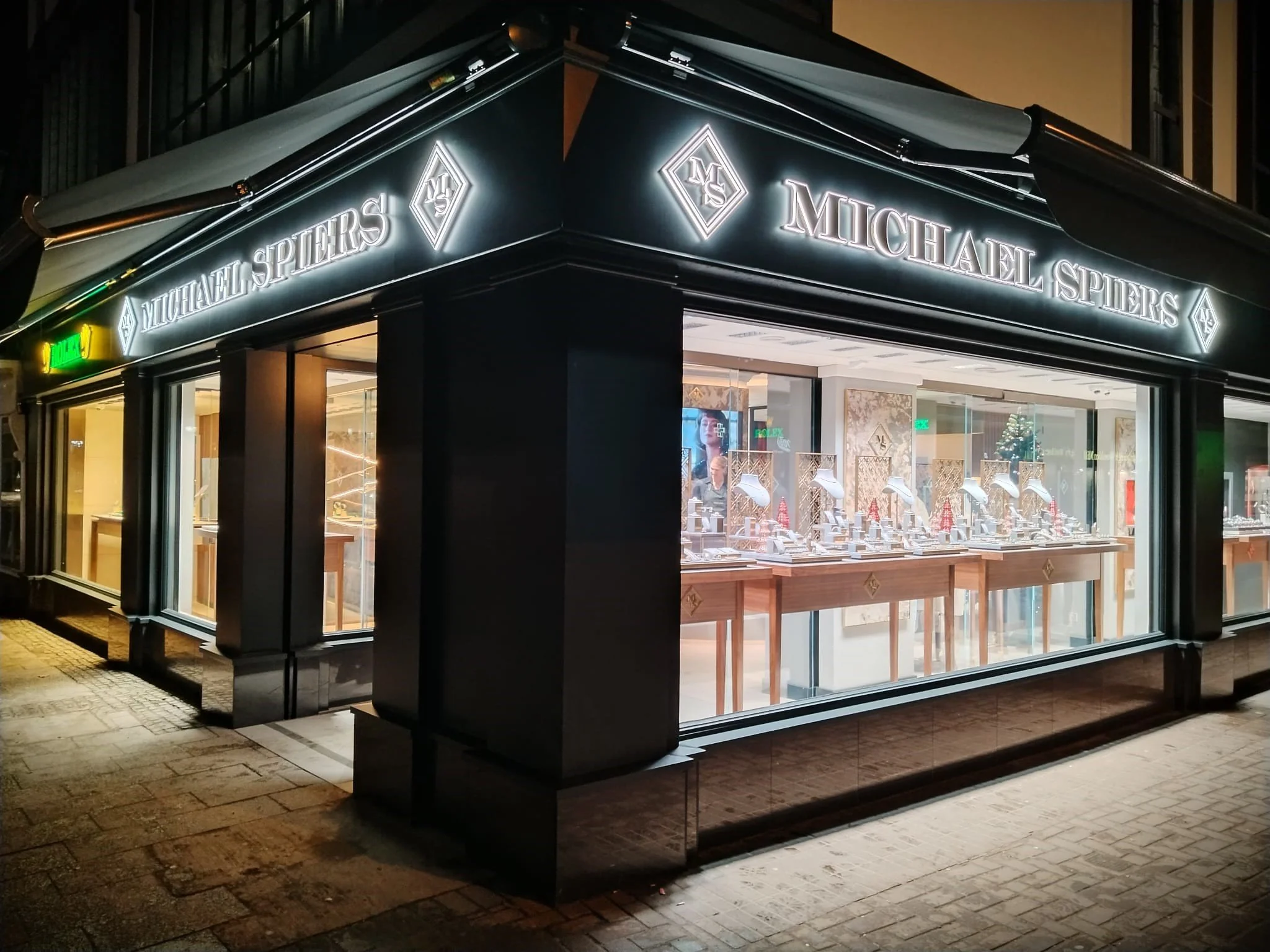 Exterior view of a Michael Spiers jewelry store at night, with large glass windows displaying jewelry and illuminated signage.
