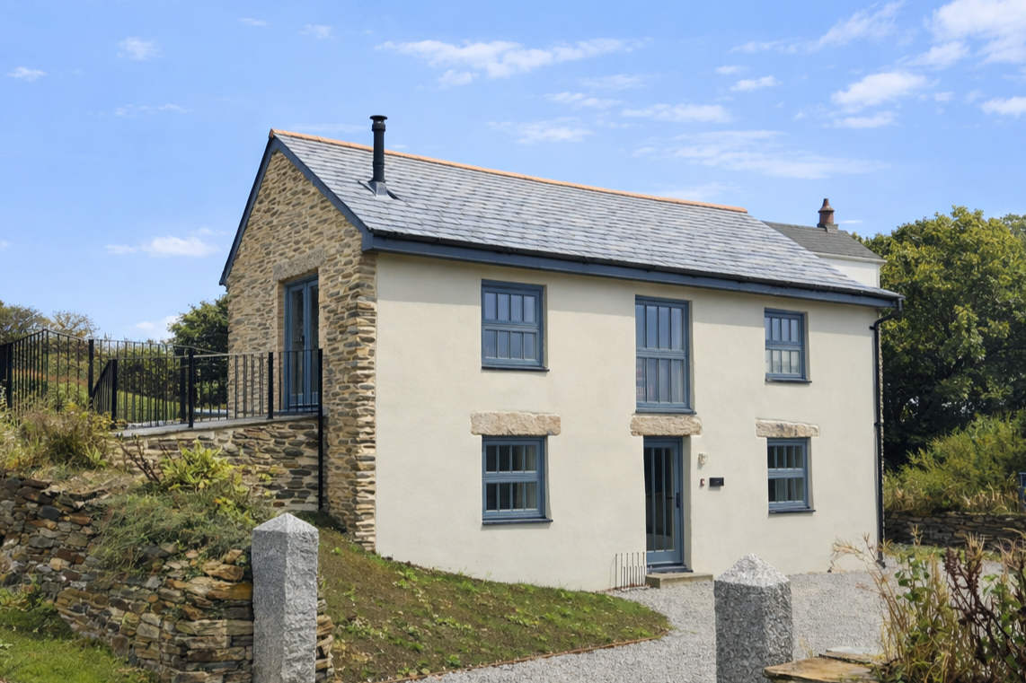 A two-story house in Cornwall with a stone and plaster exterior, blue window frames, and a gray tiled roof under a partly cloudy sky.