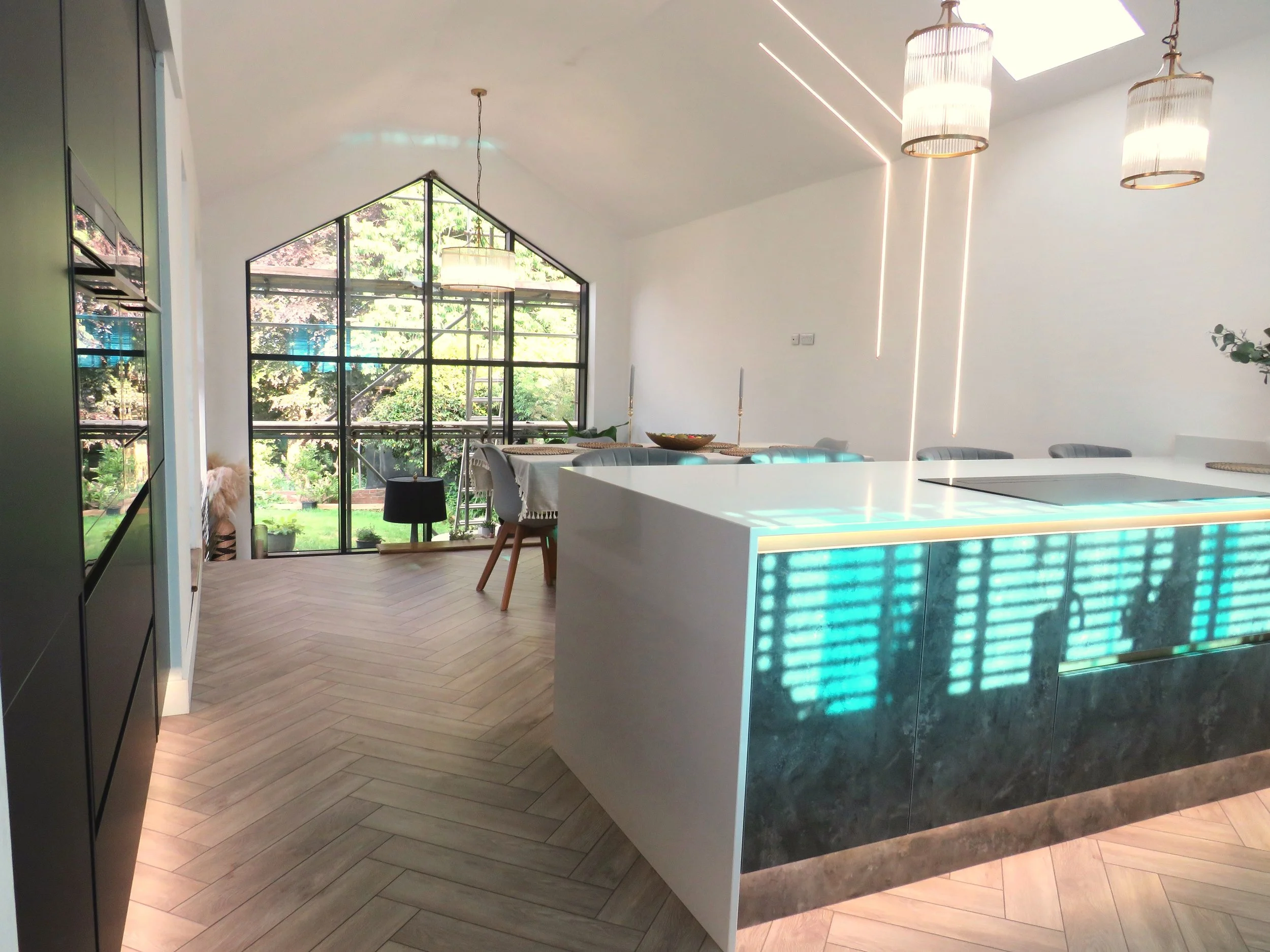 Modern kitchen and dining area with large geometric window, white walls, pendant lights, and a sleek island with built-in cooktop and illuminated front panel.