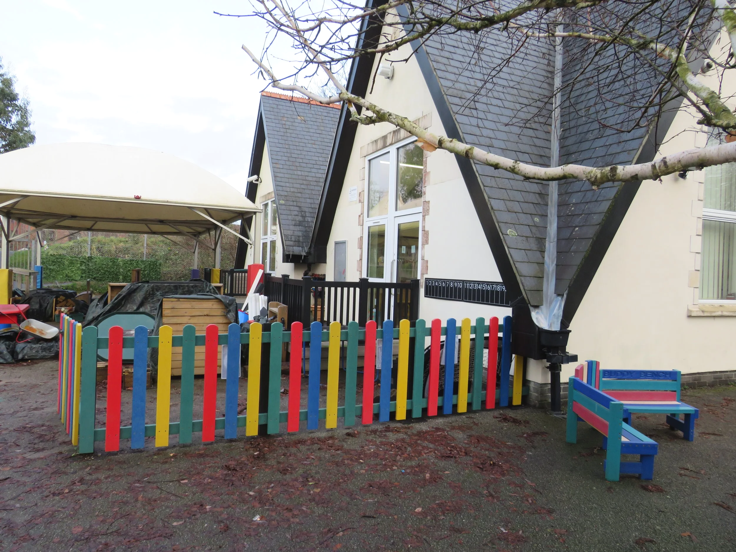 Colorful children's playground with a small painted bench, a multicolored fence, and a shaded area with toys and equipment in front of a house with hanging branches and a roof.