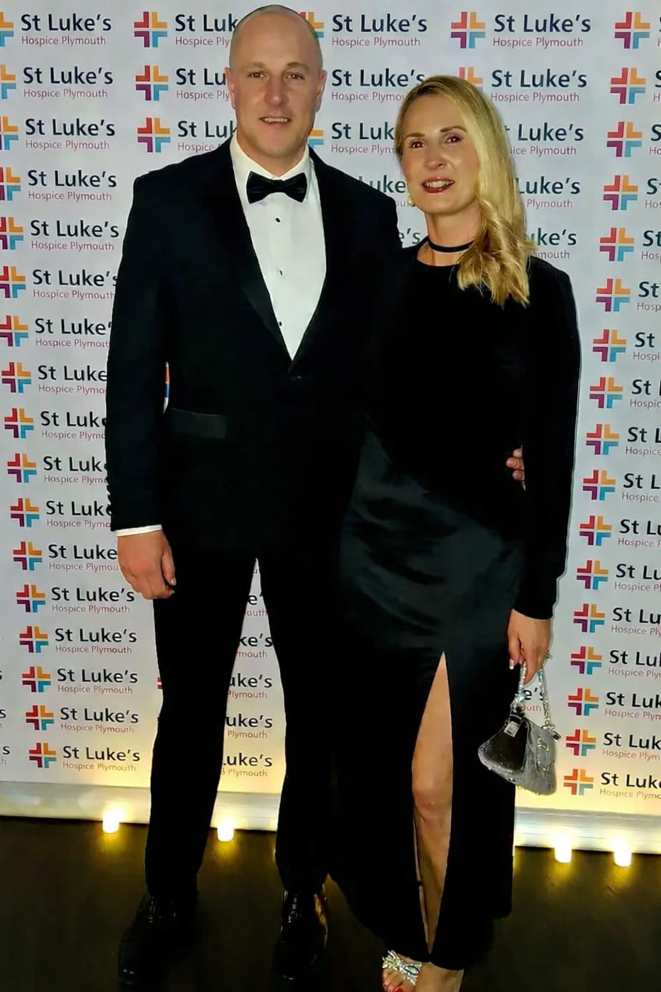 A man and woman dressed formally, standing in front of a backdrop with the Logo and text for St Luke’s Hospice Plymouth, smiling at the camera.