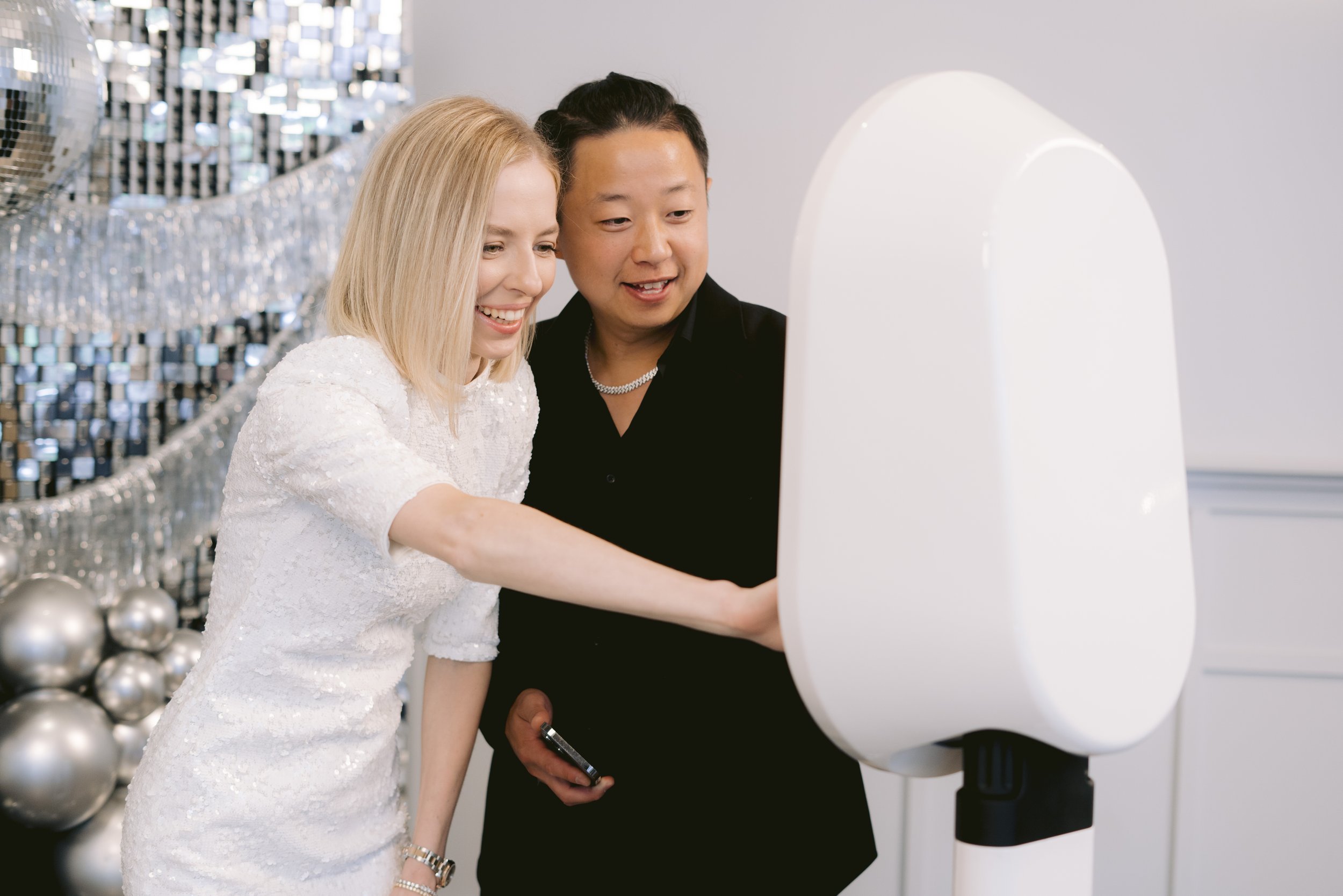 Two women smiling and laughing as they use an interactive photo booth at a celebration, with silver balloons and a mirrored background.
