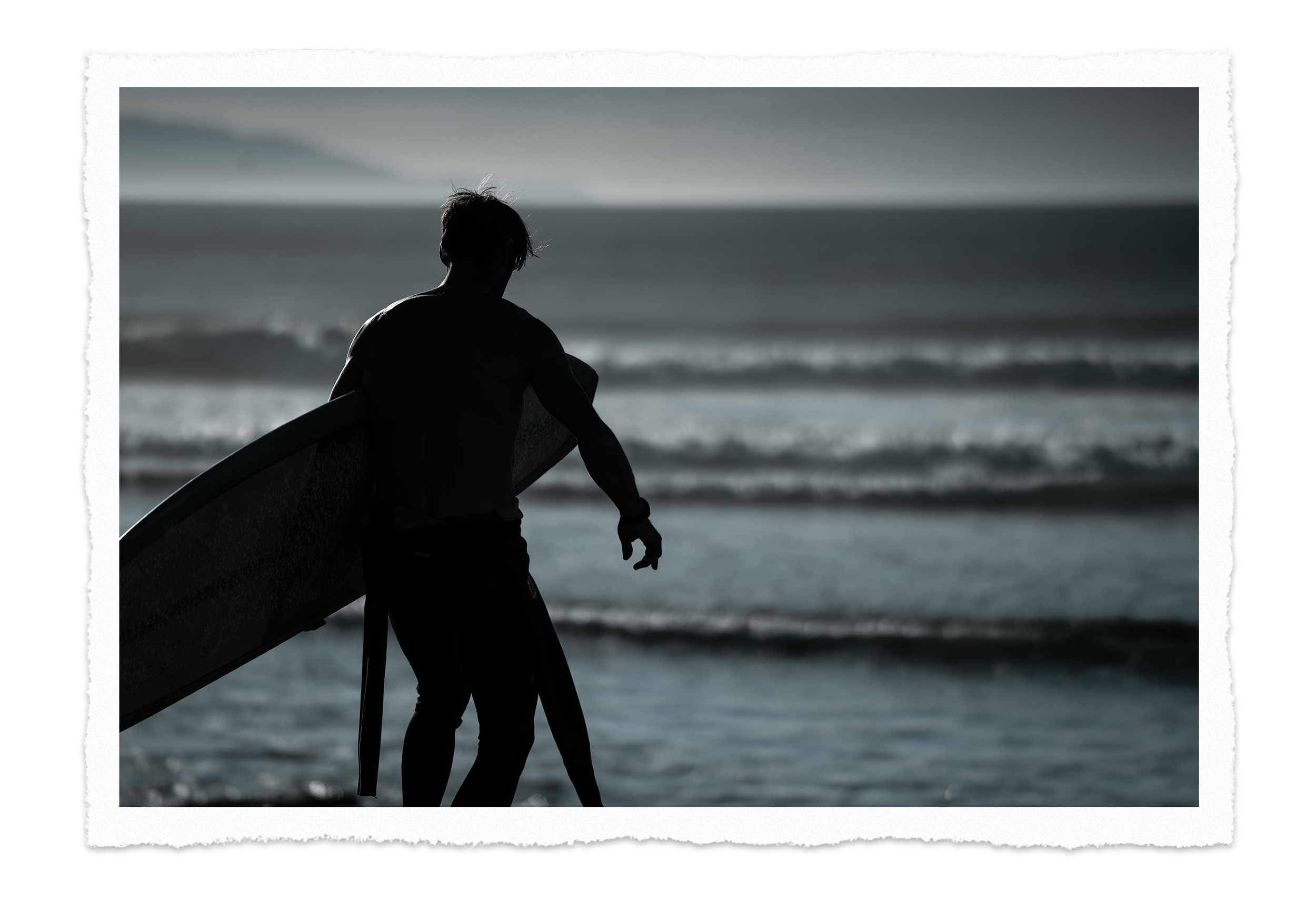 Silhouette of a male surfer holding a surfboard on a beach with waves in the background.