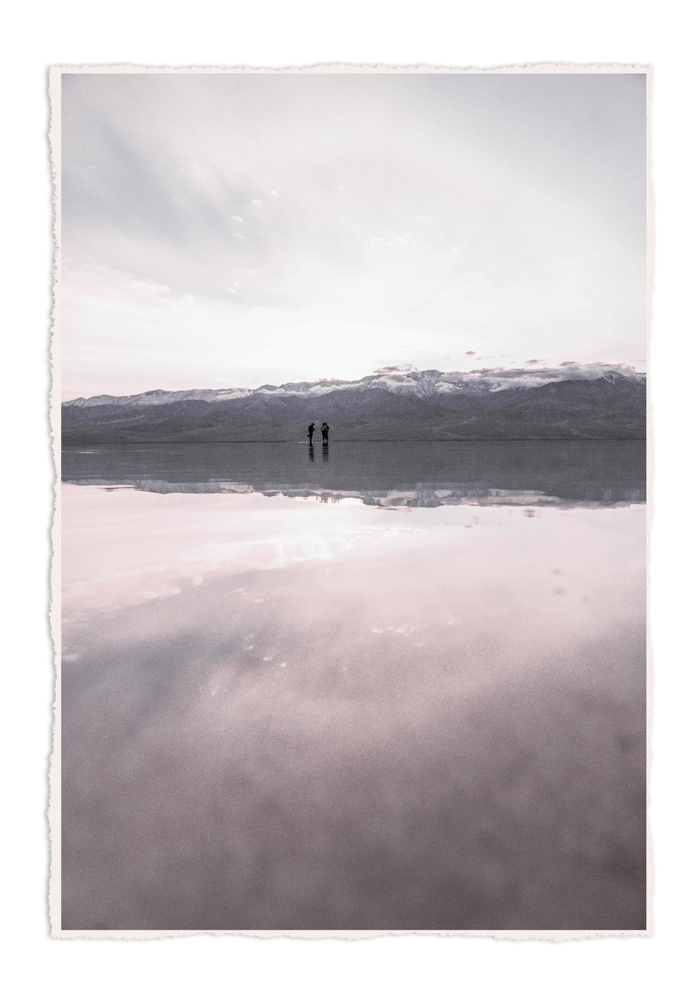 Two people standing on a reflective water surface with mountains and clouds in the background, under a cloudy sky.