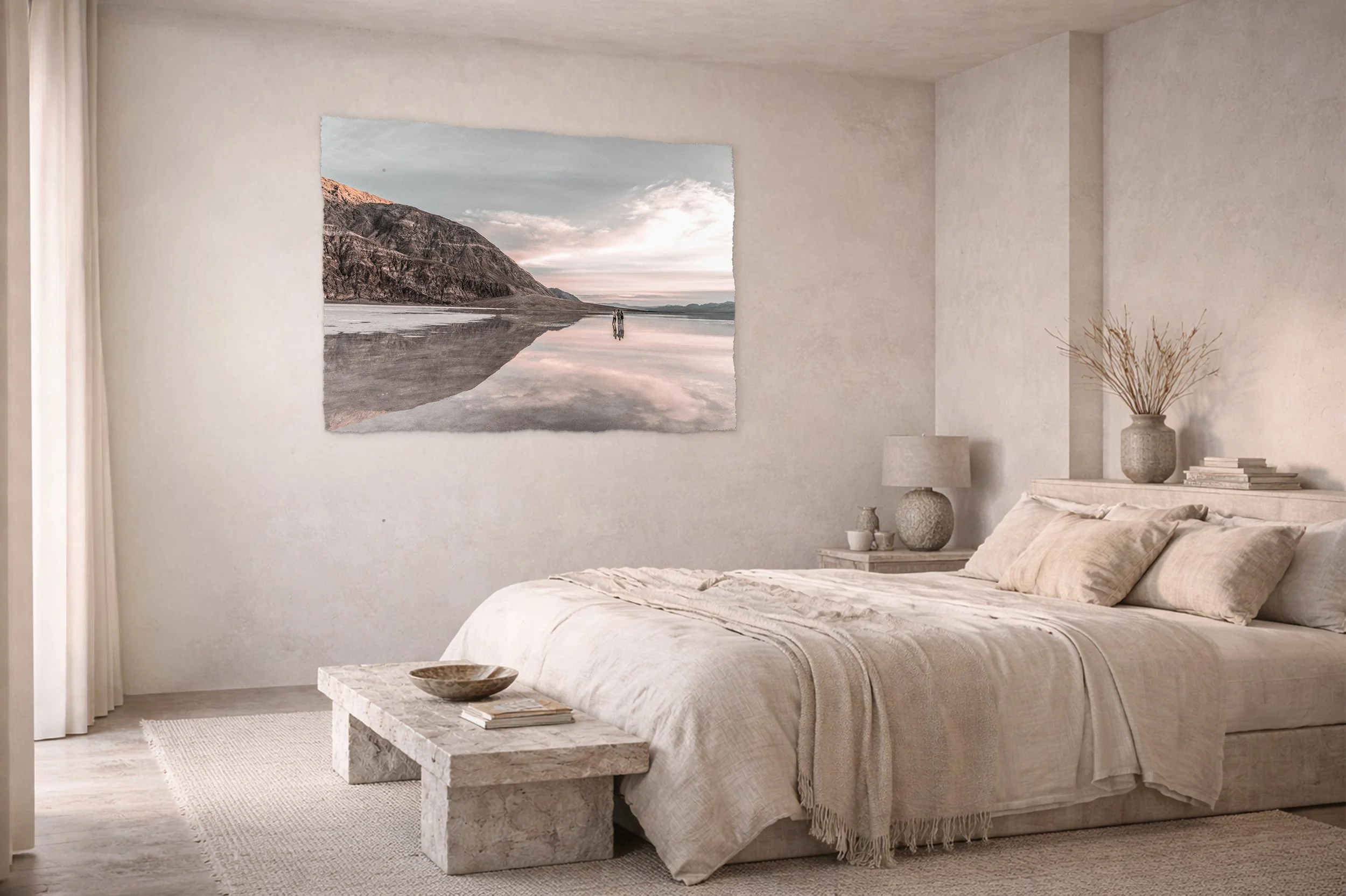 Minimalist bedroom with neutral tones, a large bed with beige bedding, a stone bedside table with a lamp and decorative items, a large landscape painting of a mountain scene on the wall, a stone bench at the foot of the bed with a bowl and books, and a window with white curtains.