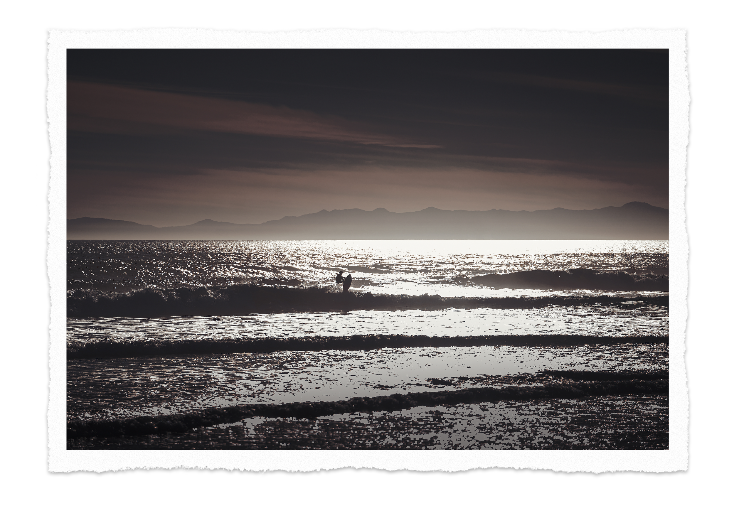 A person standing in the ocean with a surfboard in the water during sunset or sunrise, mountains in the background, and the sky partially cloudy.