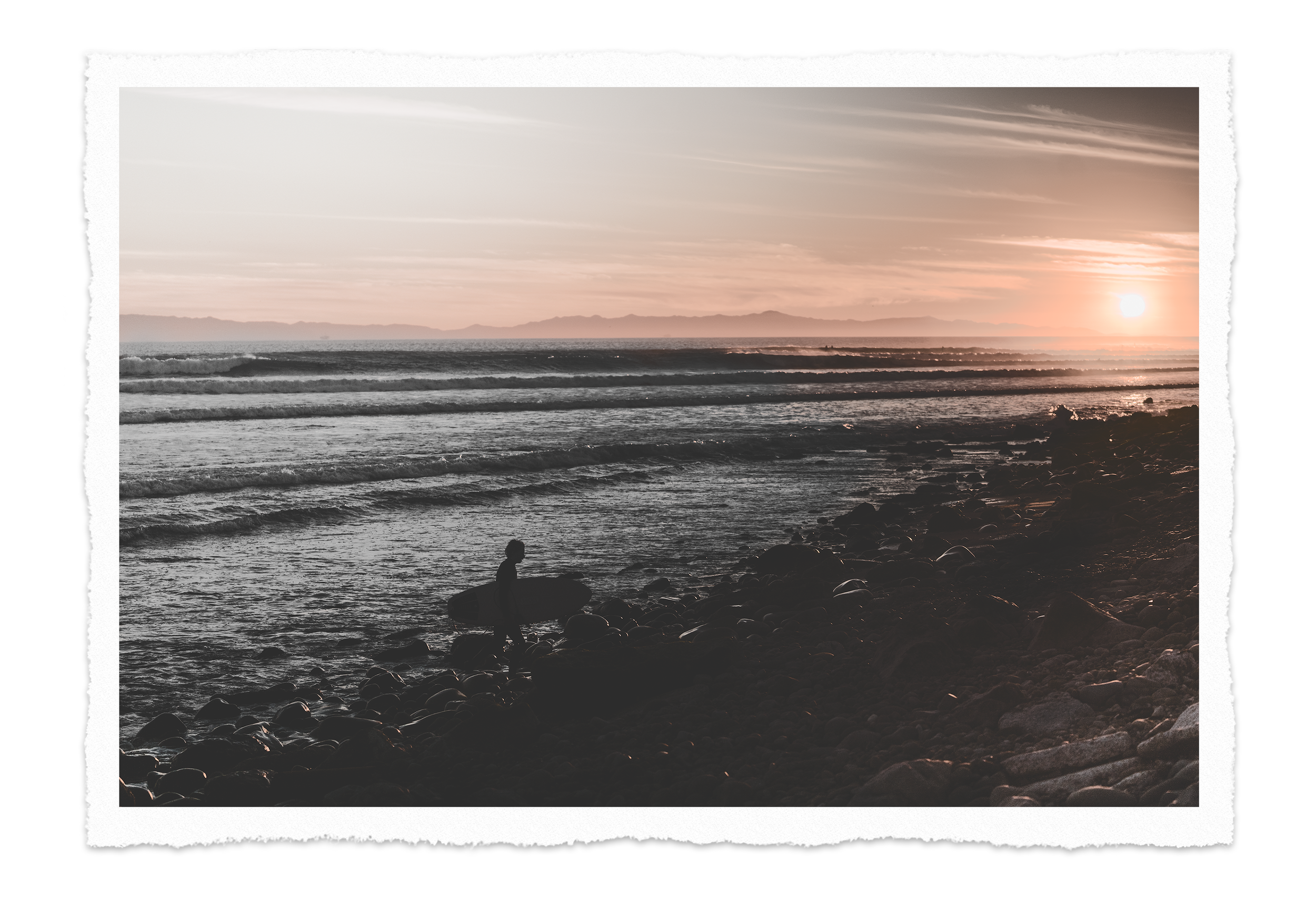 A silhouette of a person with a surfboard standing on rocks along a rocky beach at sunset, with the ocean and distant mountains in the background, in black and white with a sepia tone on the horizon.