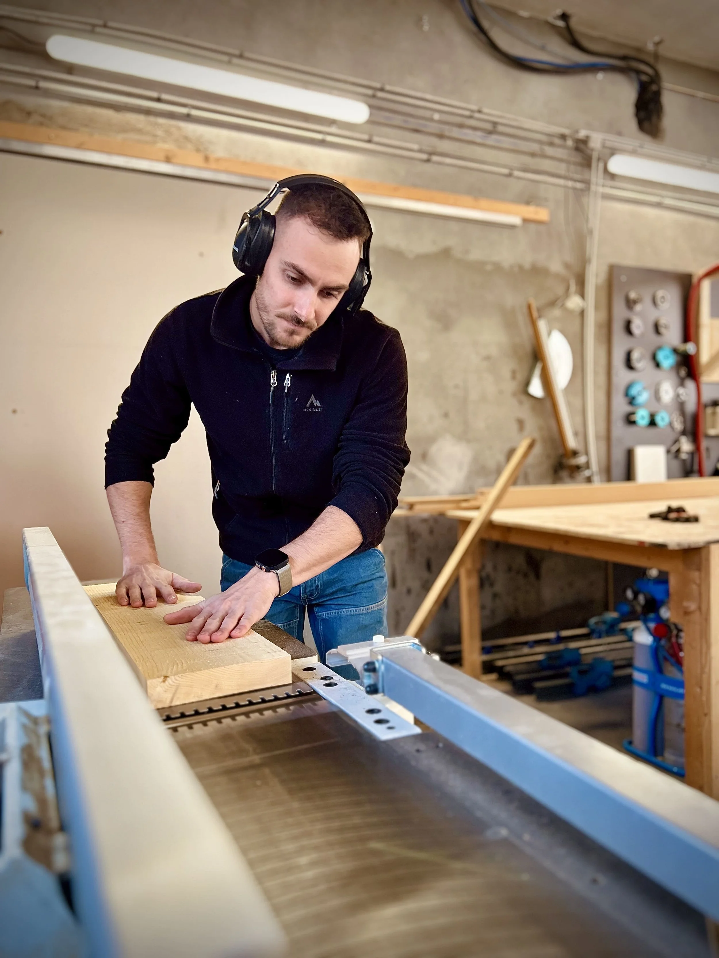 Un homme dans un atelier de menuiserie portant un casque écouteur, travaillant sur une pièce de bois avec une machine à bois.