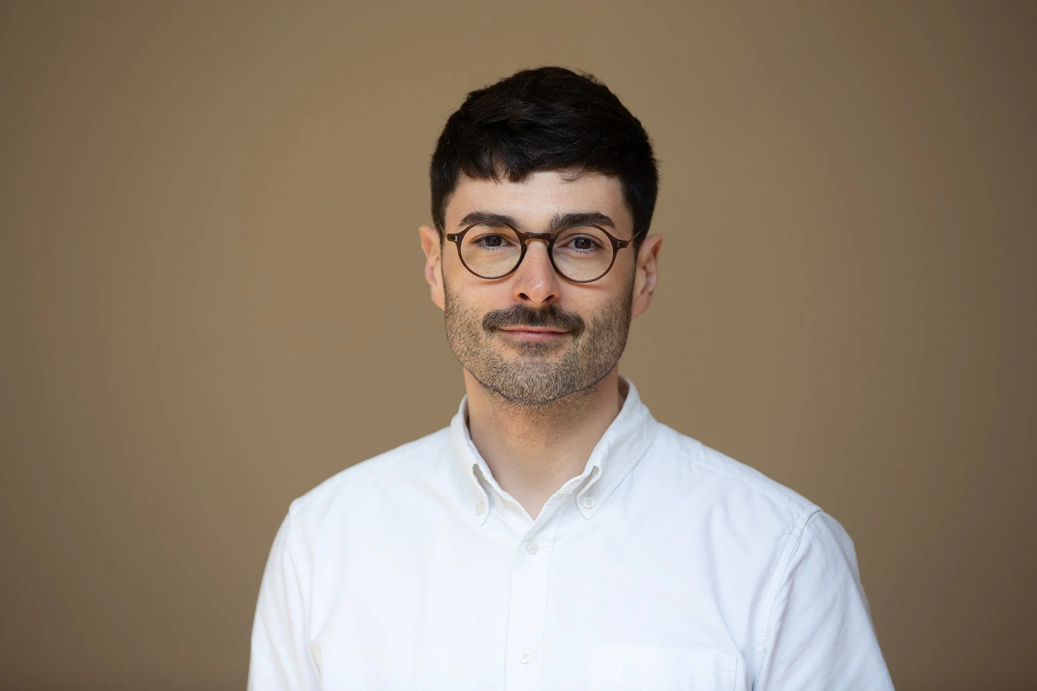 A man with dark hair, beard, and round glasses wearing a white collared shirt against a plain beige background.