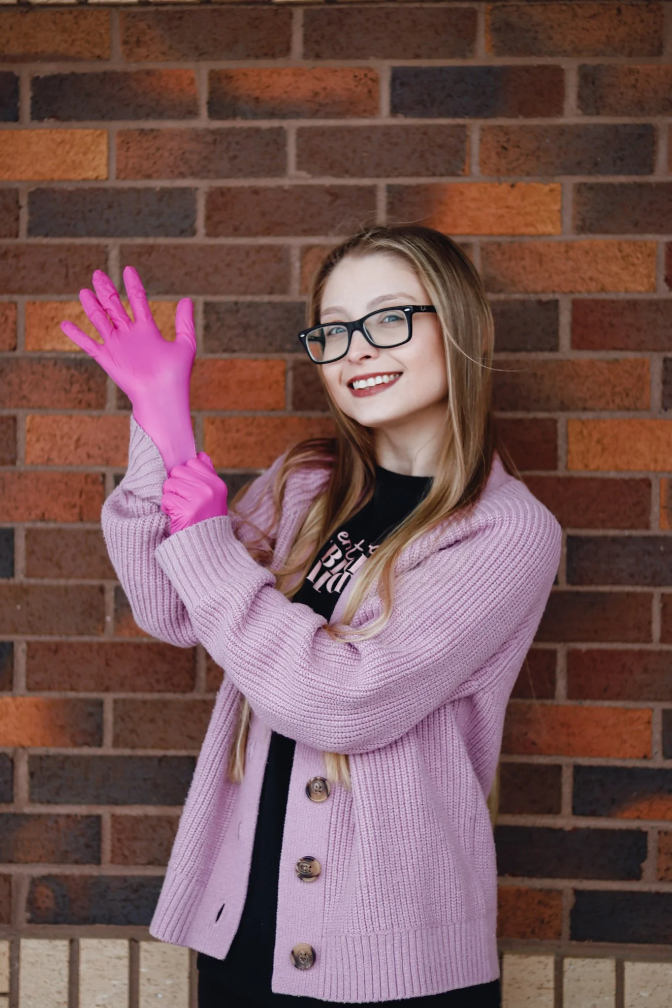 A young woman with glasses, long hair, and a pink cardigan, smiling and holding up pink rubber gloves in front of a brick wall.