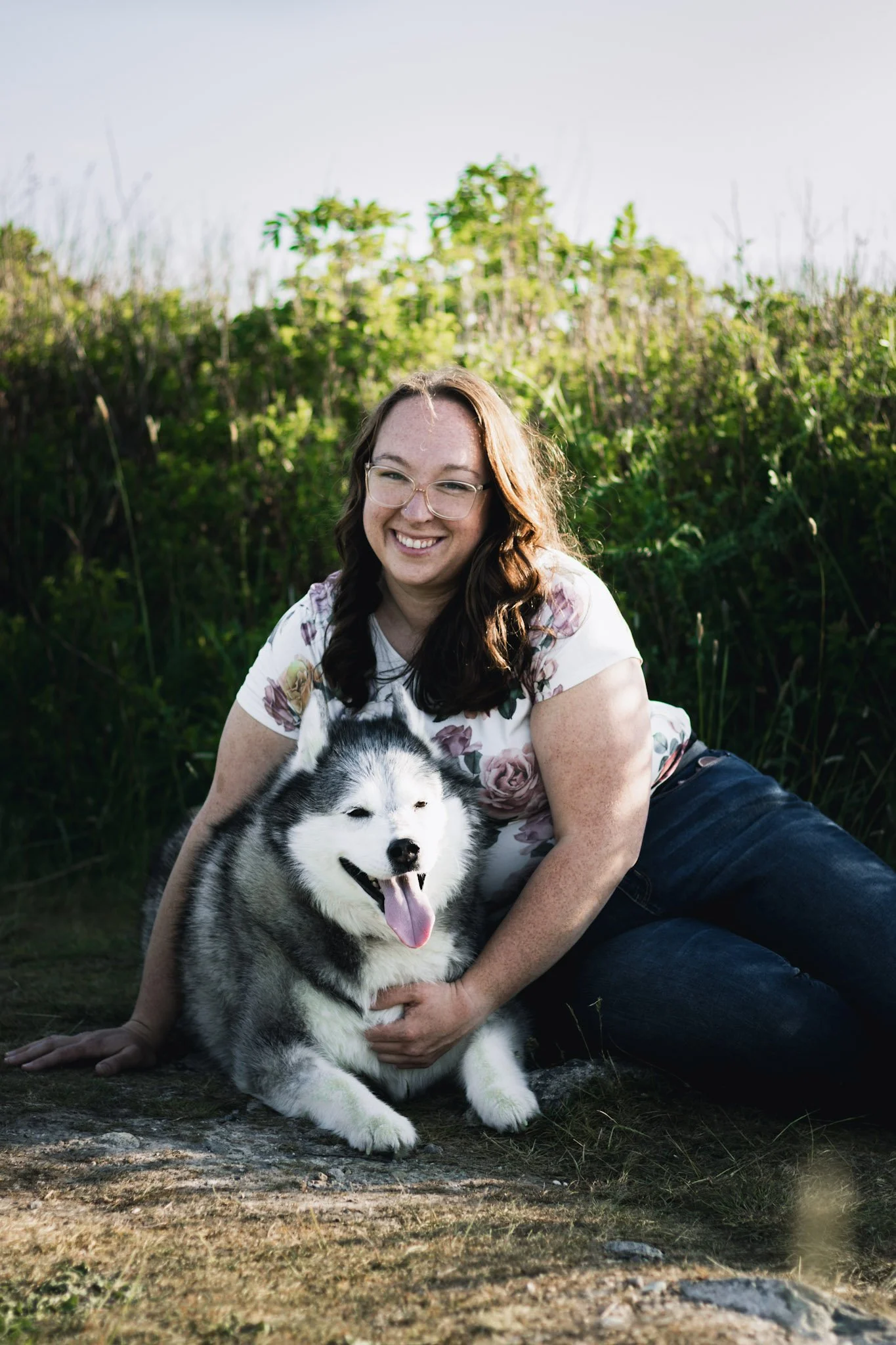 A woman with glasses and brown hair sitting on the ground outdoors next to a smiling husky puppy with black and white fur.