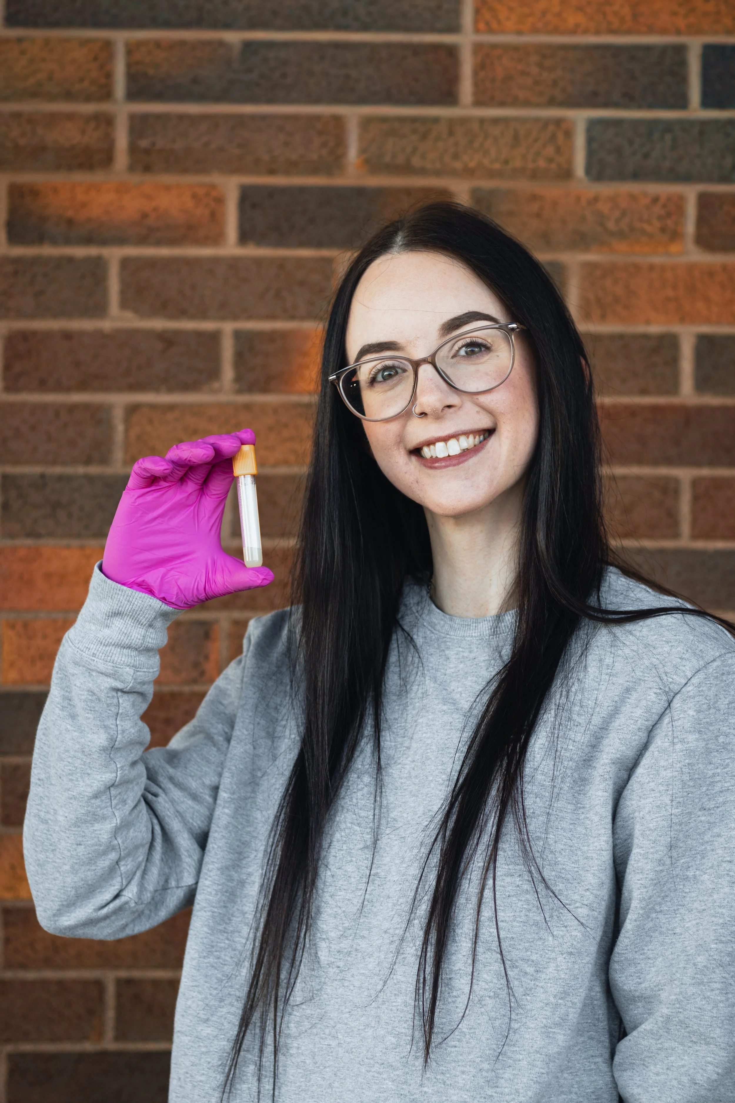 Young woman with glasses and a nose ring holding a test tube with a sample in front of a brick wall.