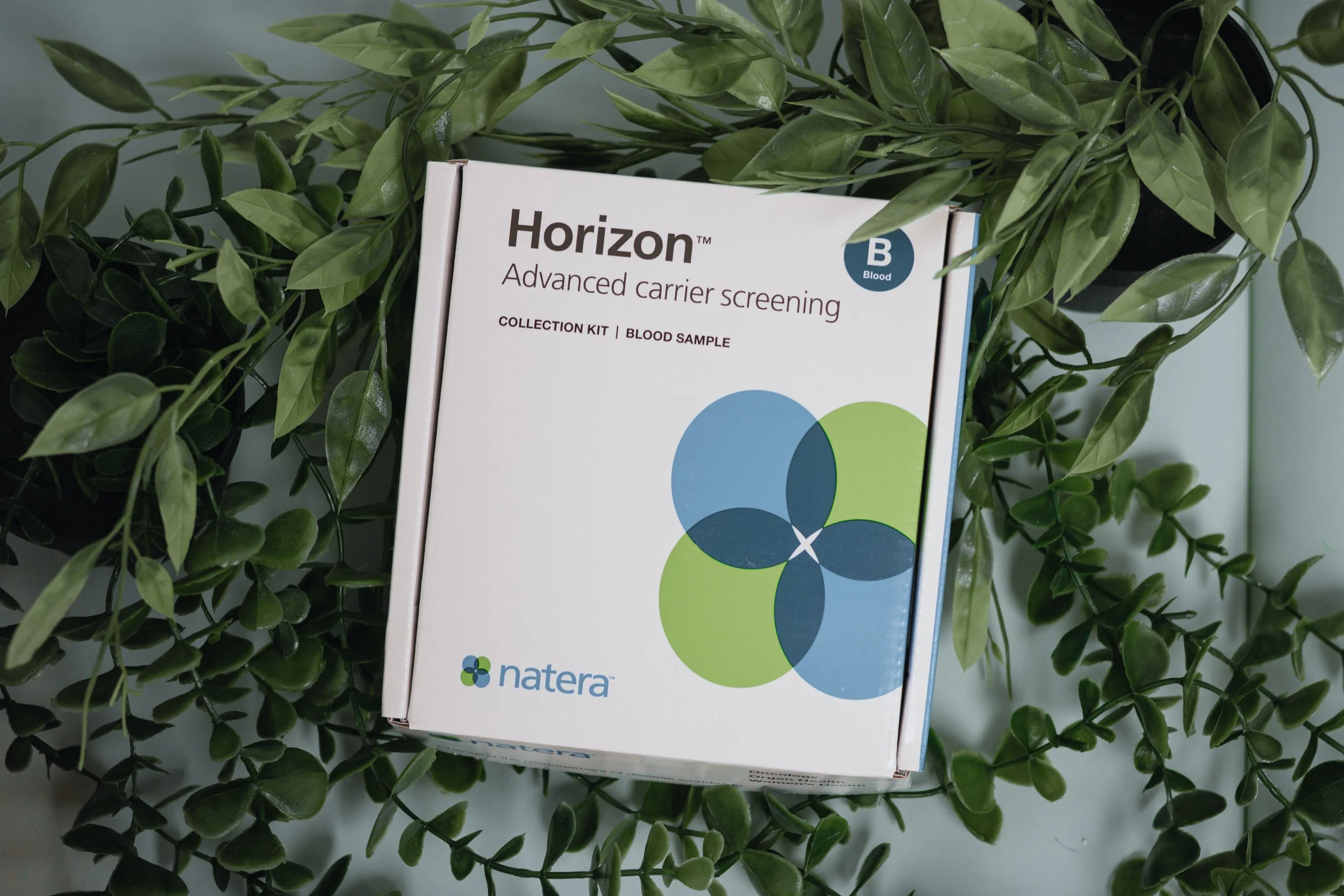 A box of Horizon advanced carrier screening blood sample collection kit on a surface surrounded by green leafy plants.