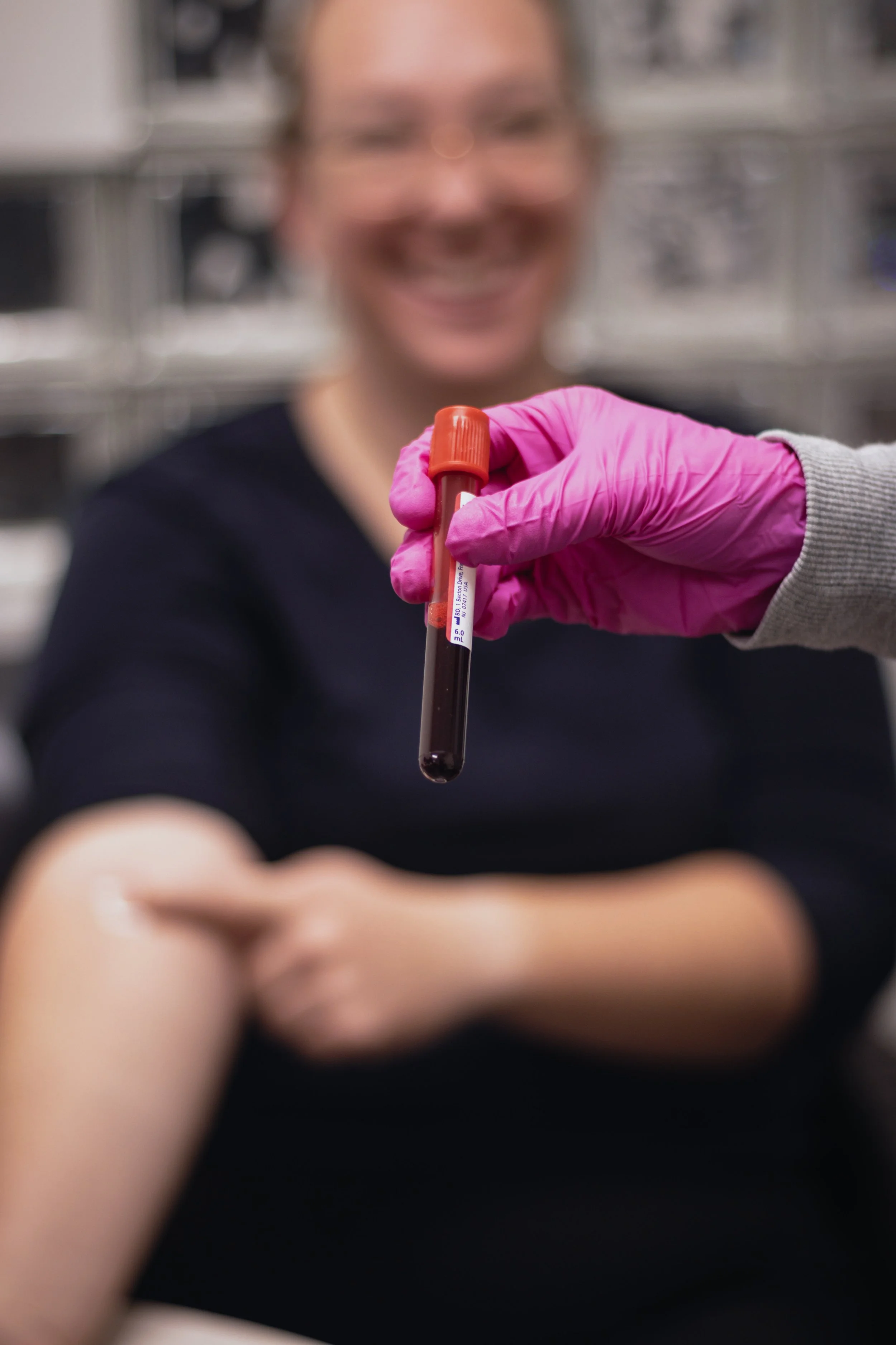 A healthcare worker in pink gloves holding a blood sample in a test tube with a smiling patient in the background.