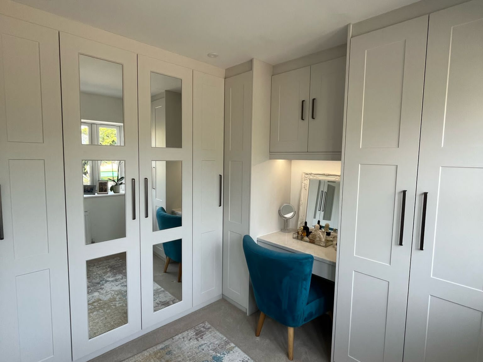 Built-in shaker wardrobes with mirrored doors and a fitted vanity area in a neutral bedroom by Eldwick Interiors.