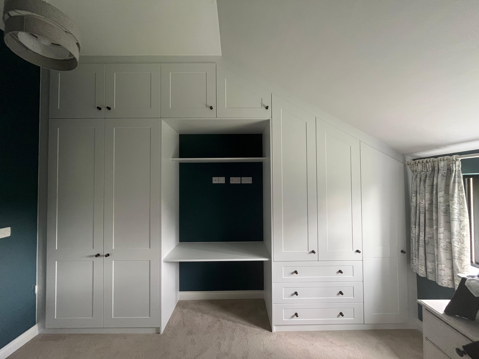 Bespoke white fitted wardrobes built into a sloped ceiling, featuring drawers, shelving and a recessed media space by Eldwick Interiors.