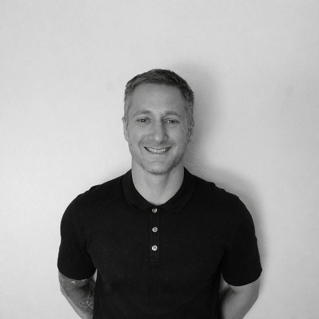 Photo en Noir et Blanc d'un homme souriant, portant un polo noir, debout devant un mur blanc.