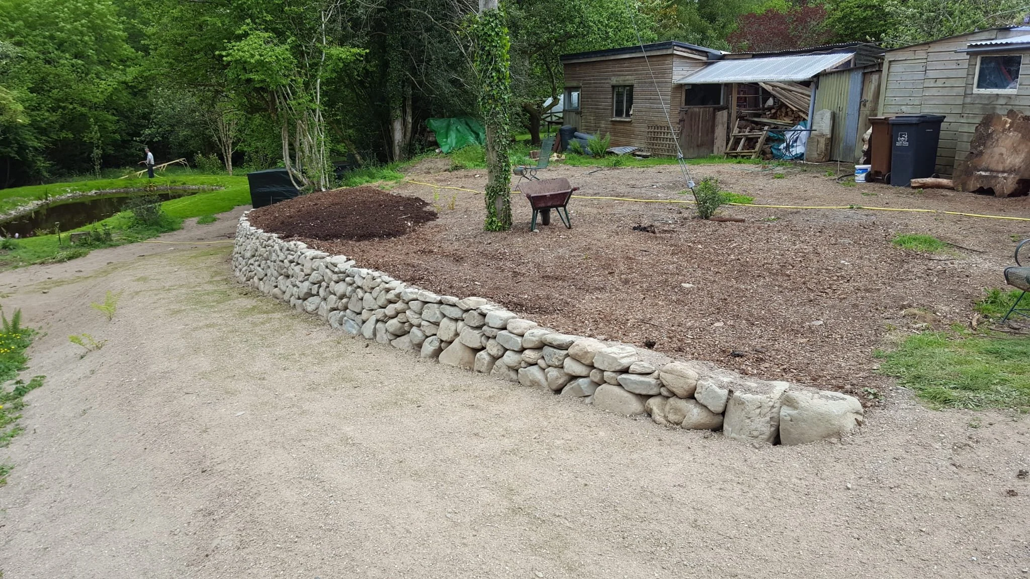Dry stone retaining wall, trees, a small pond, garden tools, and a wooden shed.