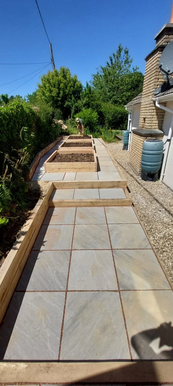 Working on building a garden pathway with large stone tiles and wooden frames for garden beds along the side in a garden under sunny weather.