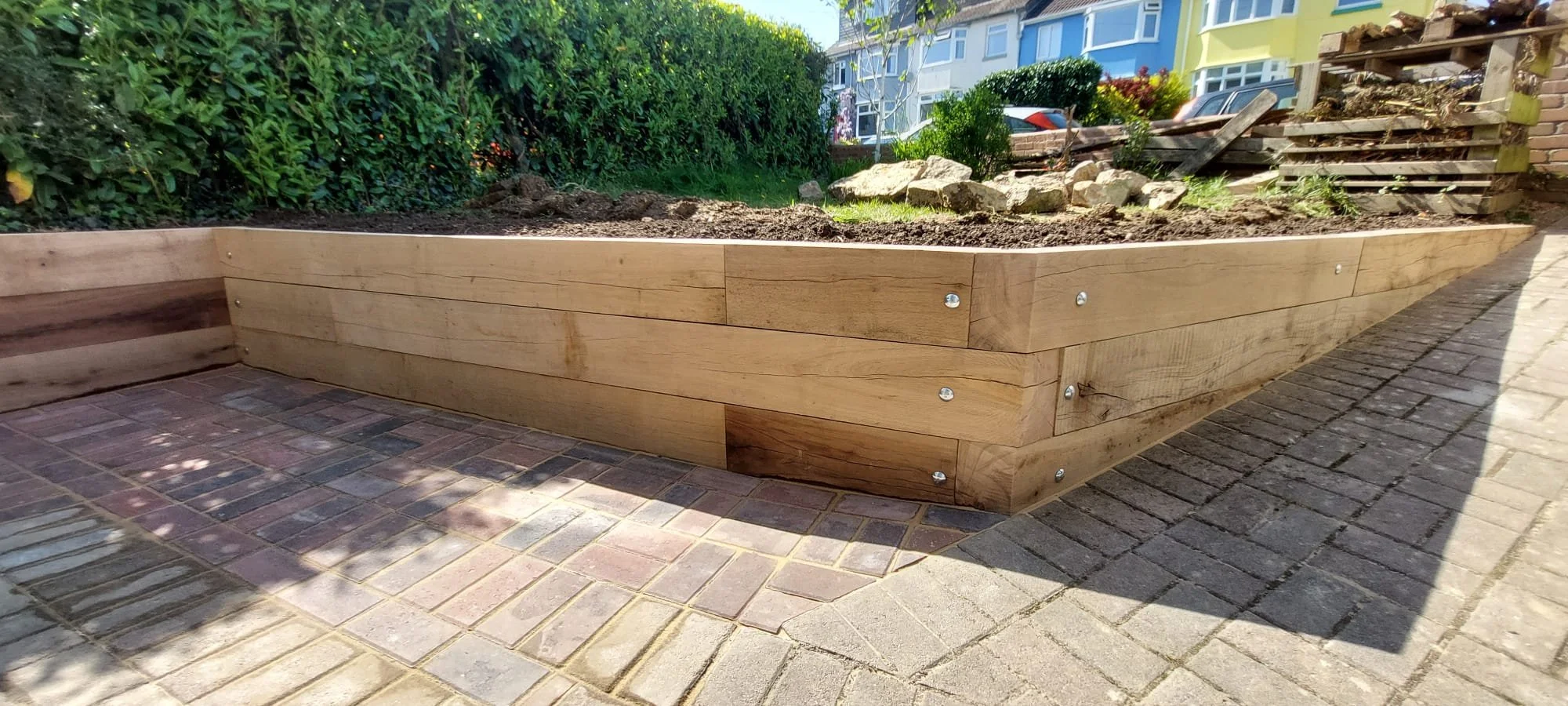 Timber retaining wall with plants and soil, situated on a paved patio with colourful houses in the background.