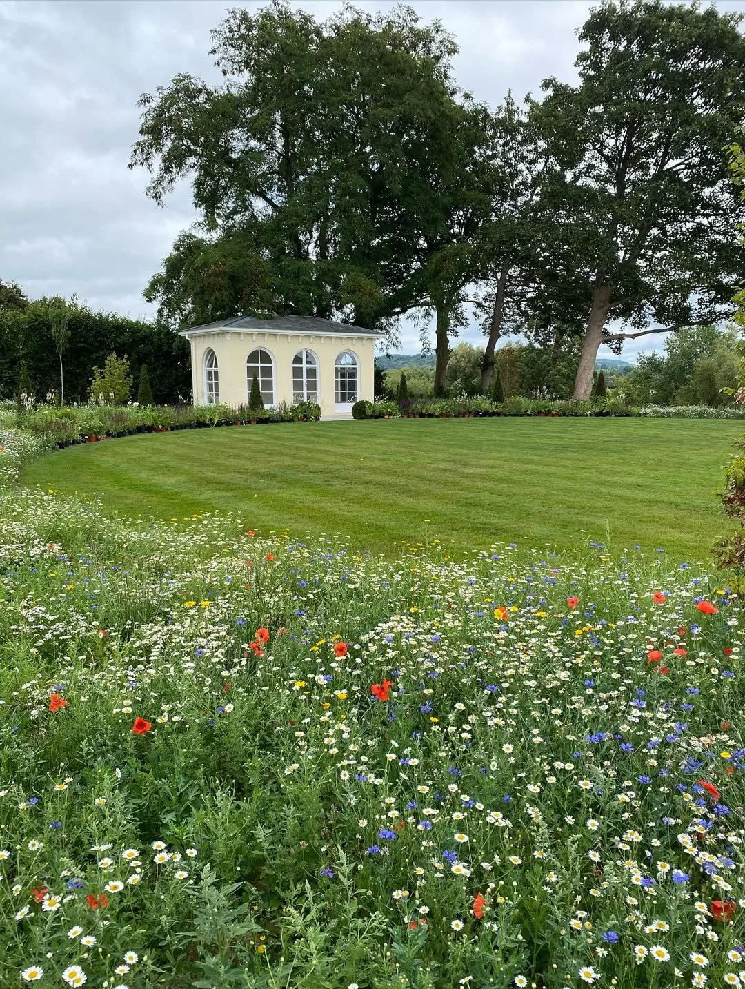 A garden with a lush green lawn, colorful wildflowers in the foreground, and a small, cream-colored pavilion with large arched windows in the background, surrounded by trees.