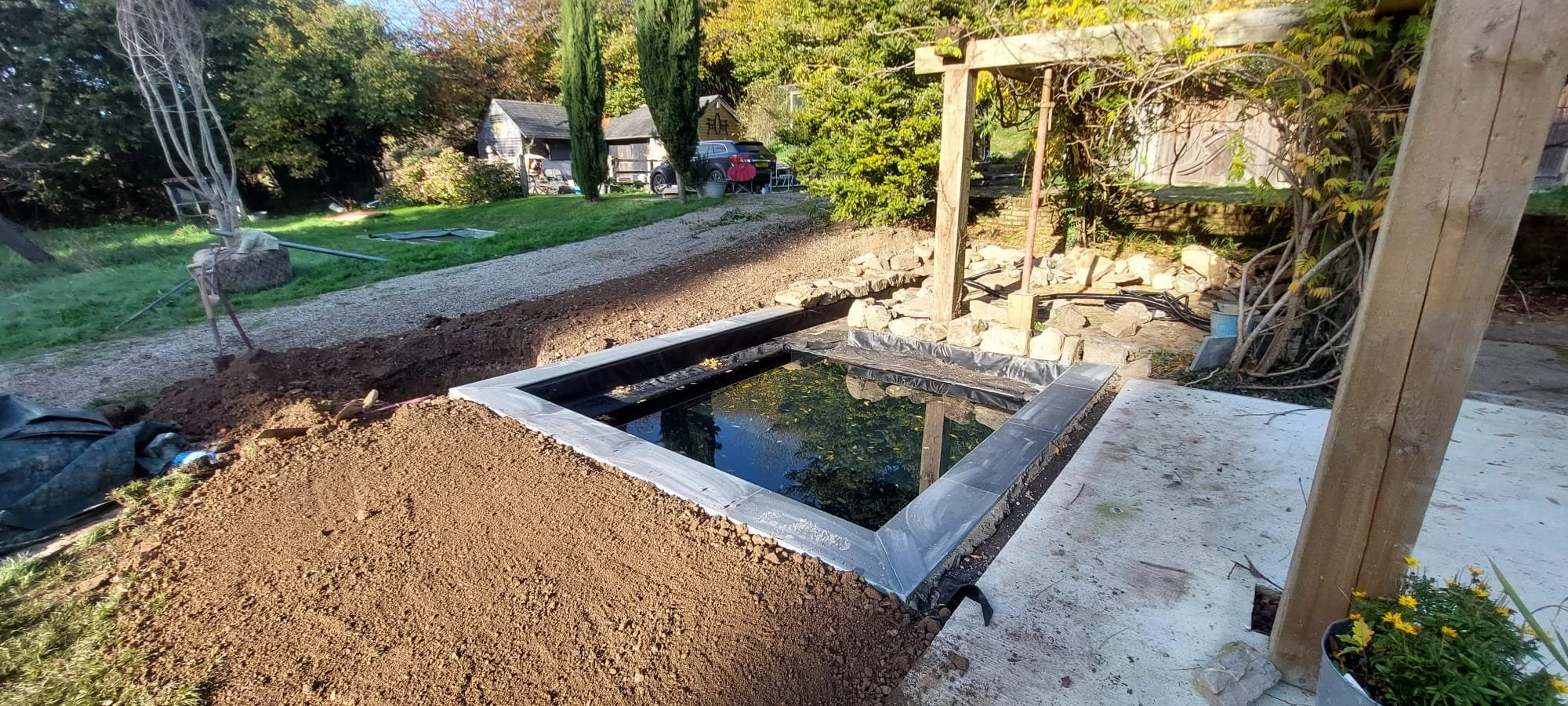 A small rectangular pond with a black liner and a metal frame, surrounded by dirt and construction materials, in a garden with trees, a gravel path, and a shed.
