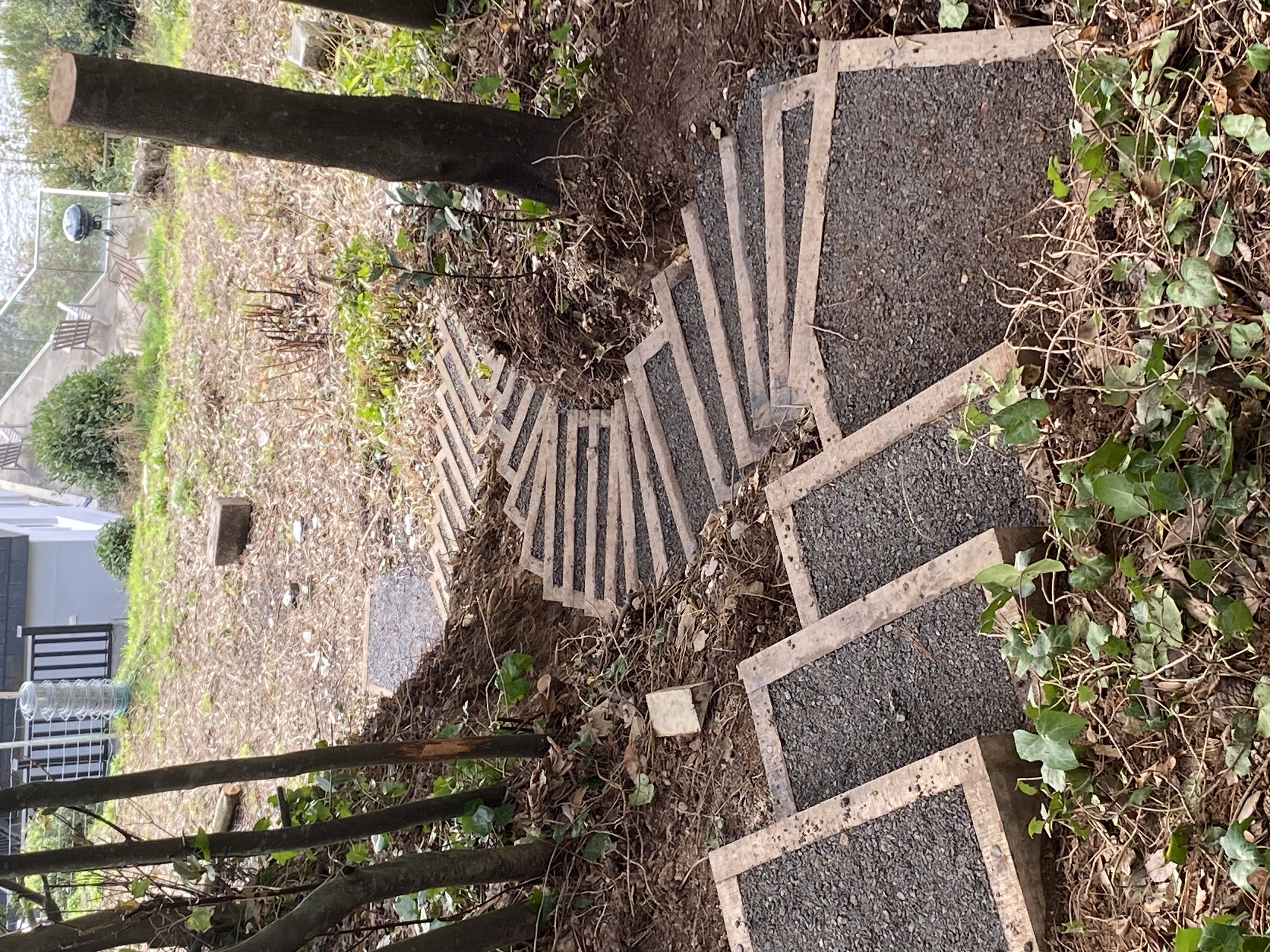 Stairs curving down a steep garden, surrounded by dirt and plants, with a tree and a balcony in the background.