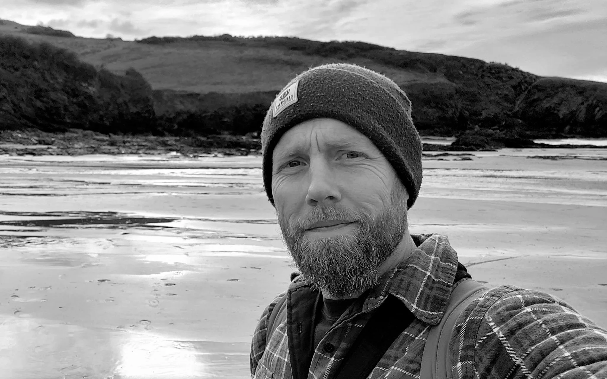 Paul taking a selfie on a beach, wearing a beanie and checkered shirt, with hills and a cloudy sky in the background.