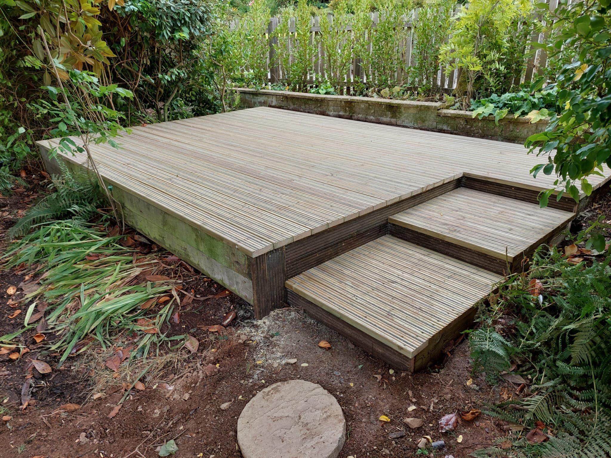 A wooden deck with two steps leading up to it, situated in a garden with greenery and plants surrounding it.